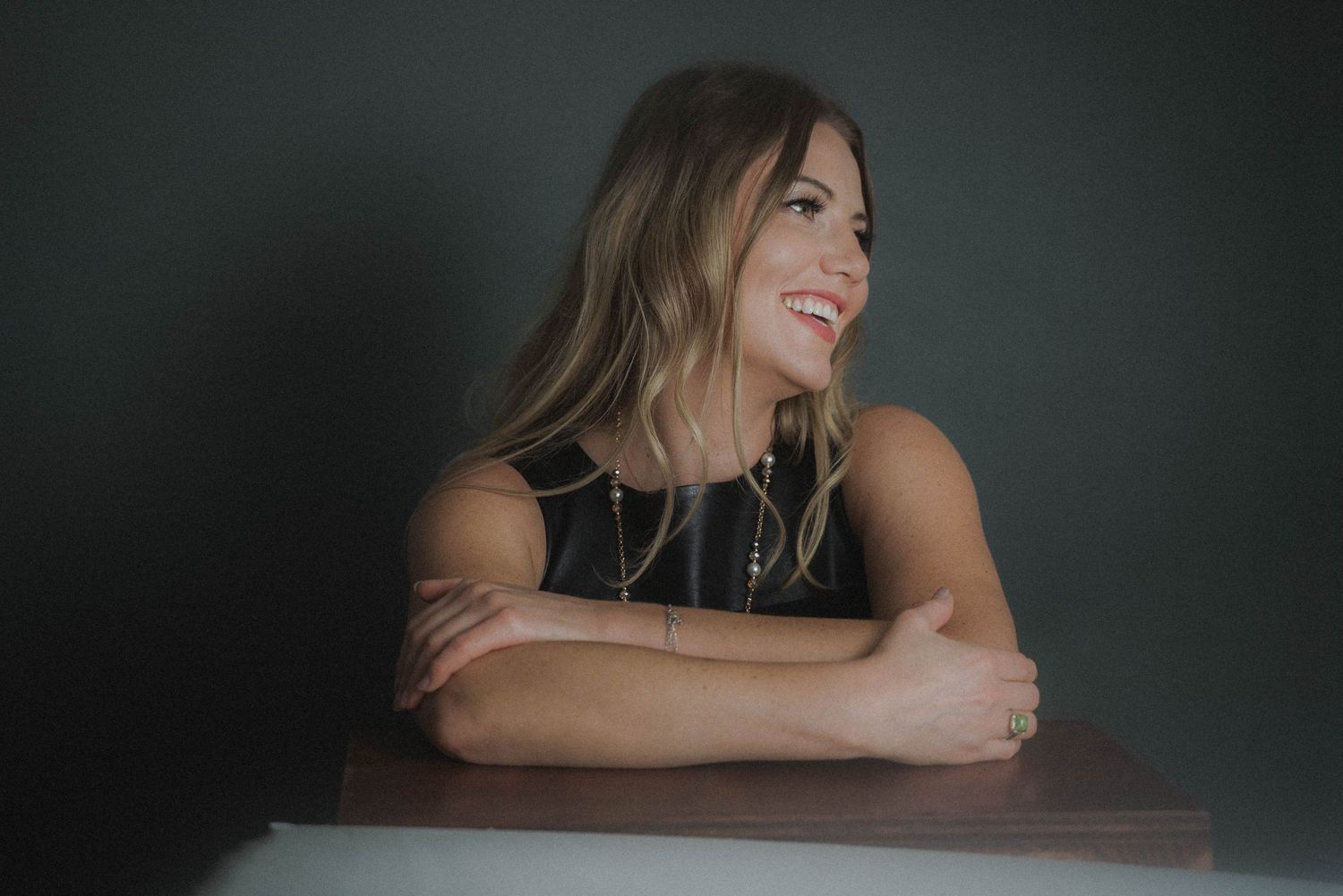 A figure sits at a table with crossed arms, wearing a black sleeveless top and smiling warmly against a dark background.