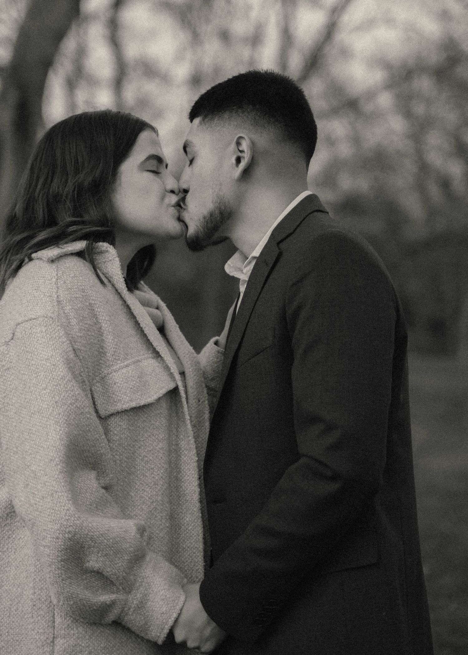 A couple shares an intimate kiss in a romantic black and white portrait outdoors with a blurred nature background.
