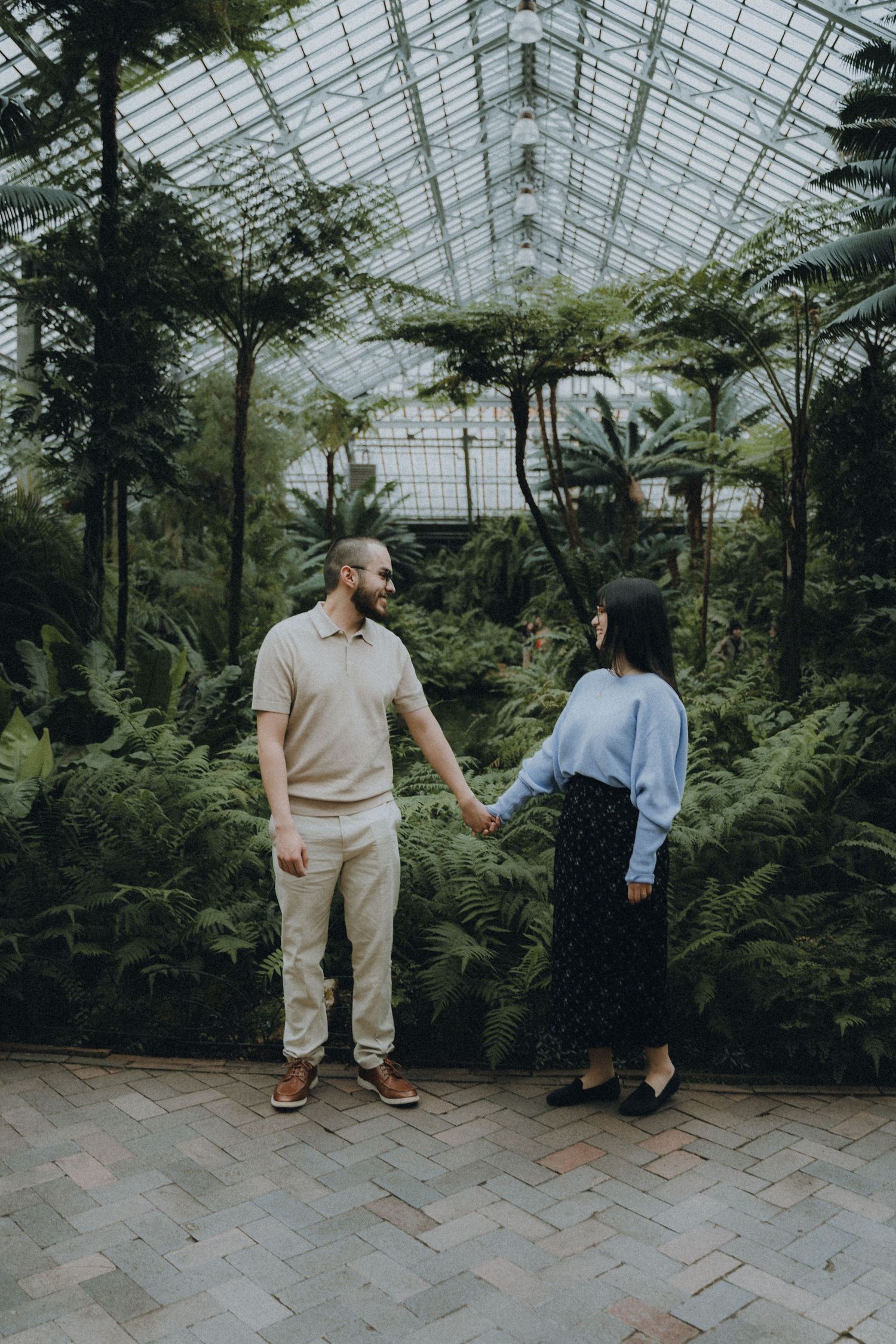 Two people stand in a greenhouse filled with lush ferns and tropical plants on a brick pathway.