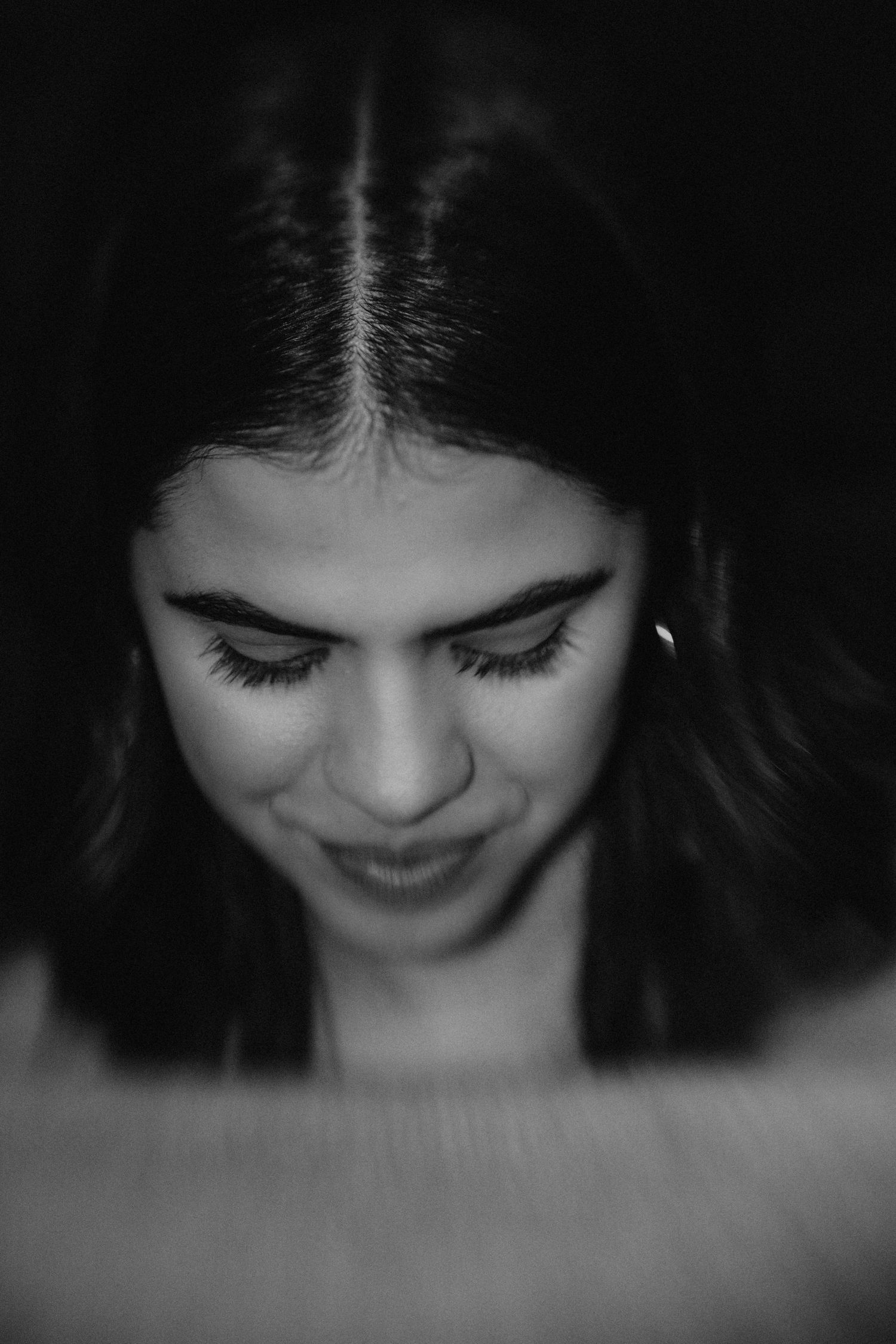 Black and white portrait of a young woman with long dark hair looking down, smiling softly in dramatic lighting.