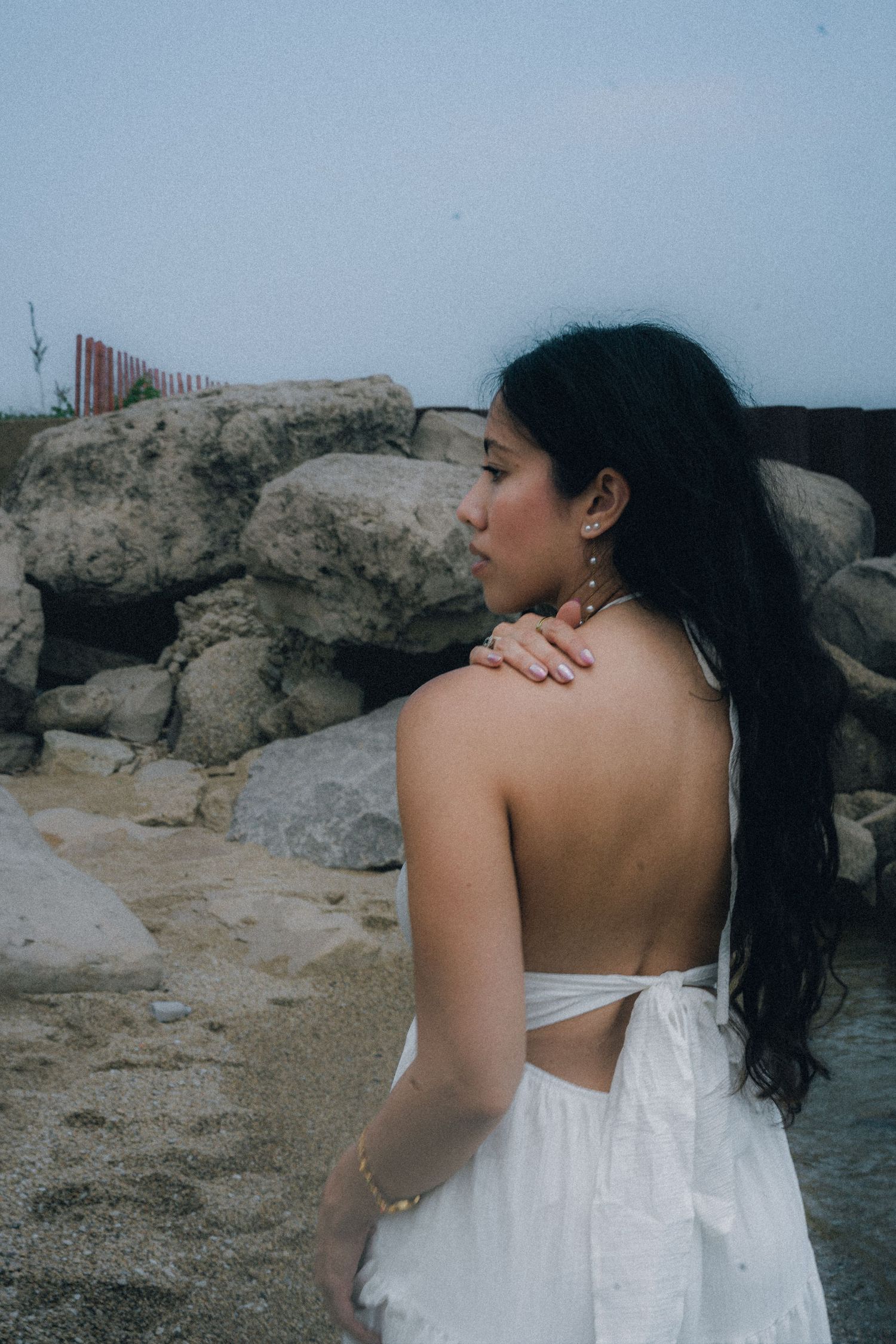 Woman in white backless dress standing near rocky beach, wearing gold earrings and bracelet.