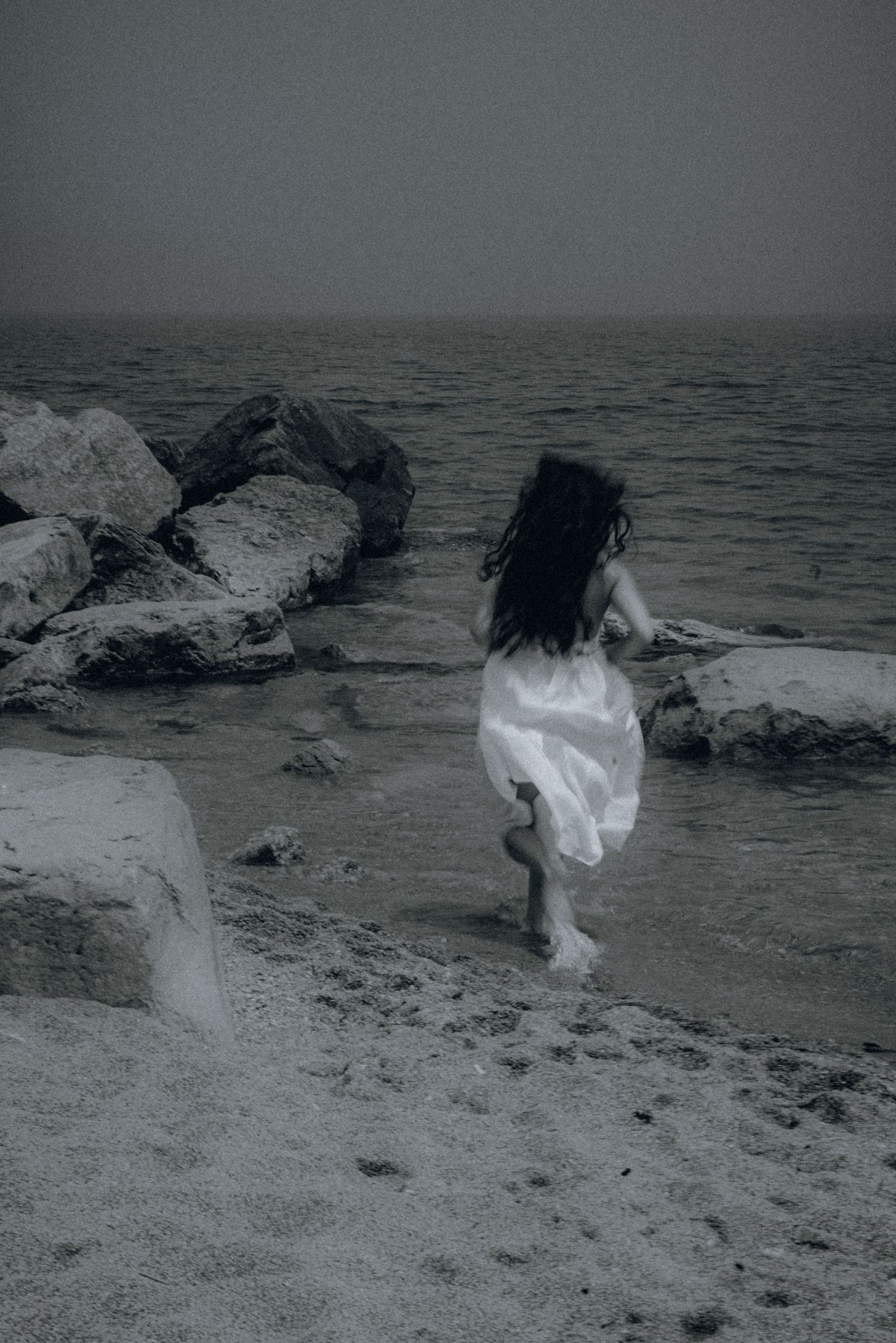 A woman in a white dress runs along a rocky shoreline in a moody black and white coastal scene.