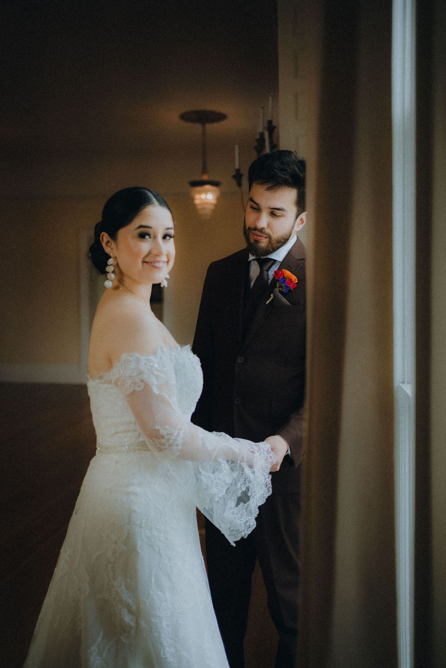 A bride in a white lace gown and groom in dark suit share intimate moments in a dimly lit hallway.