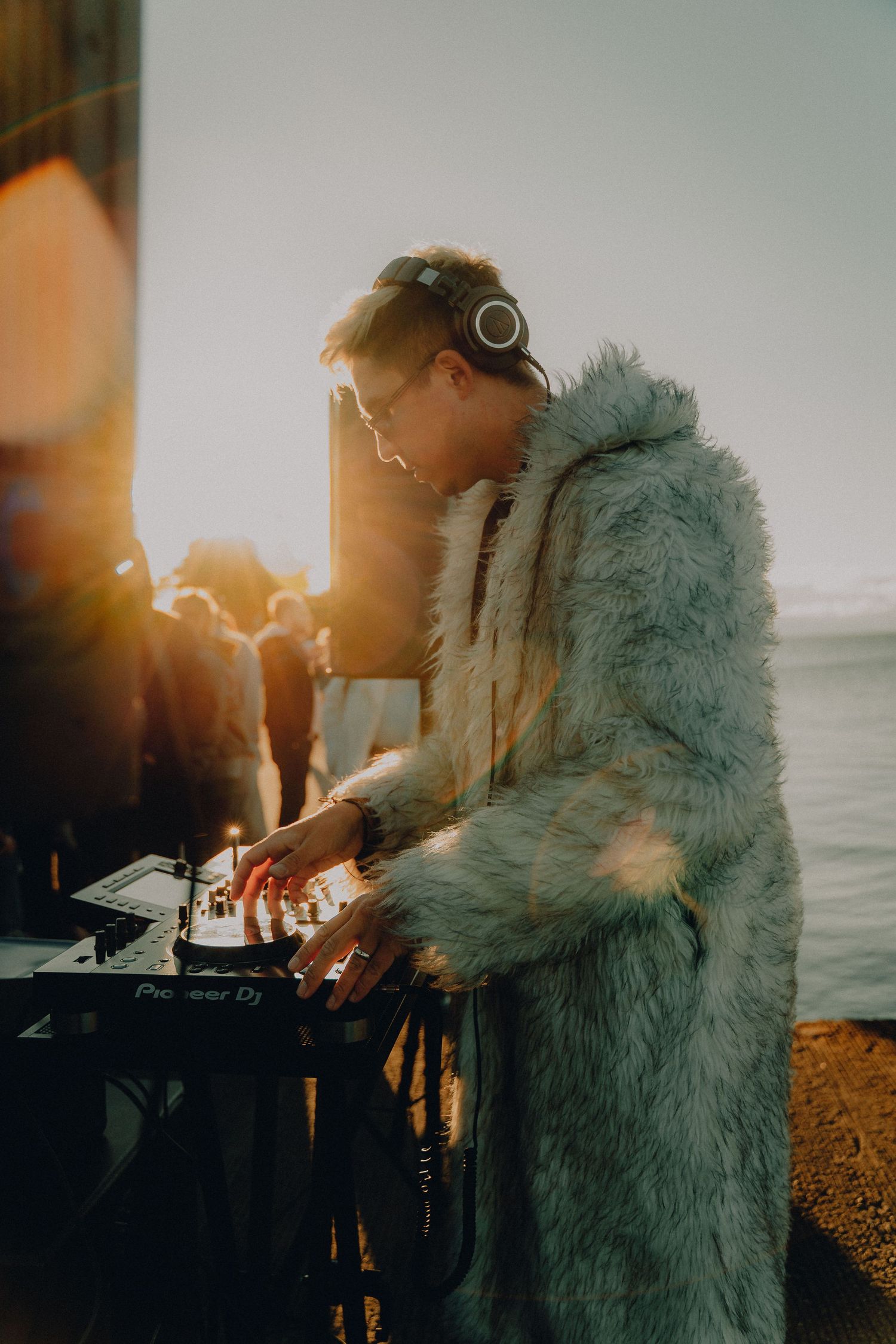 DJ wearing a fuzzy blue coat performs on turntables at sunset against an ocean backdrop.