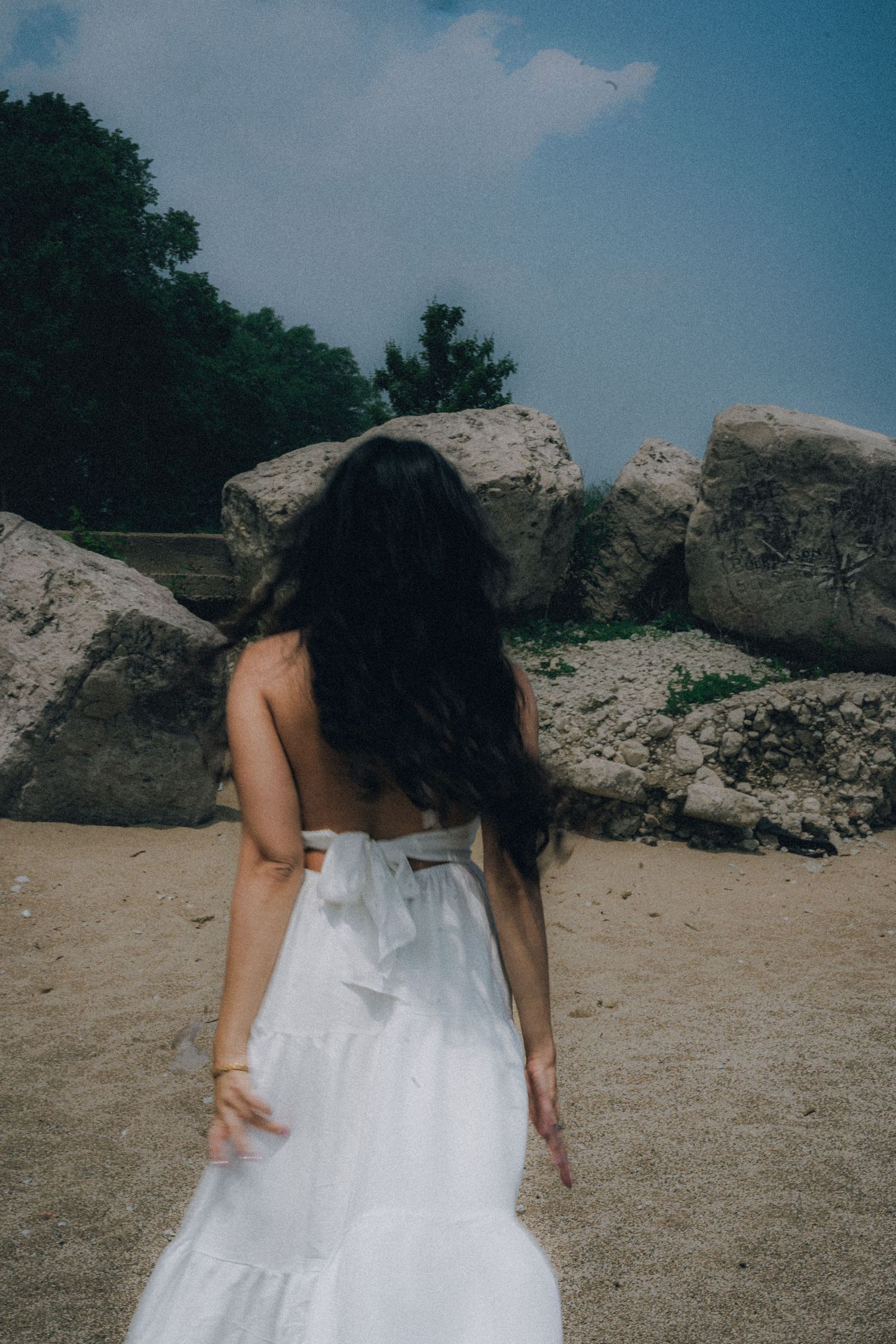 Woman in white backless dress with long dark hair walks toward large rocks on a sandy beach.