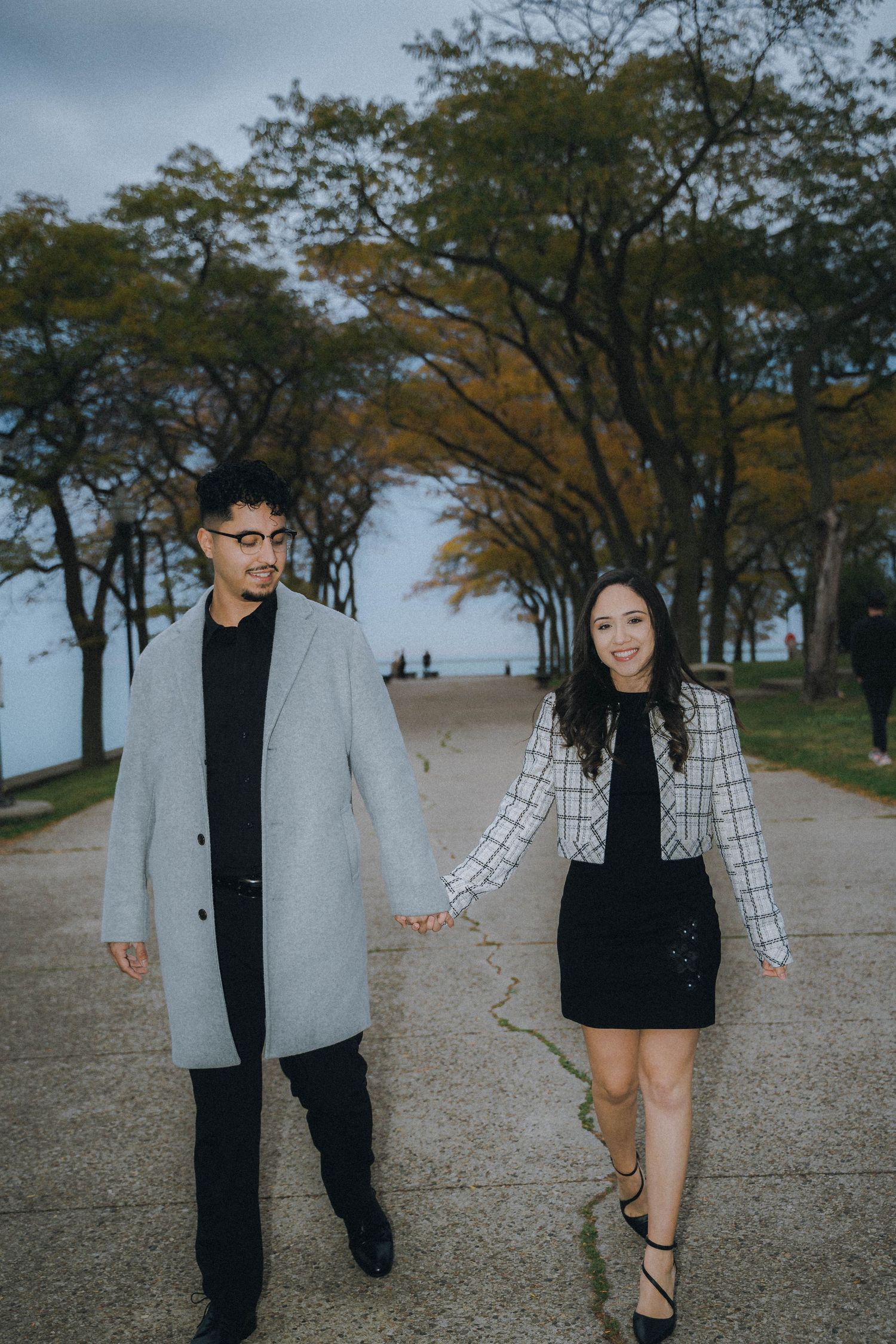 A couple walks hand in hand along a tree-lined path on an autumn day, wearing coordinated black and gray outfits.