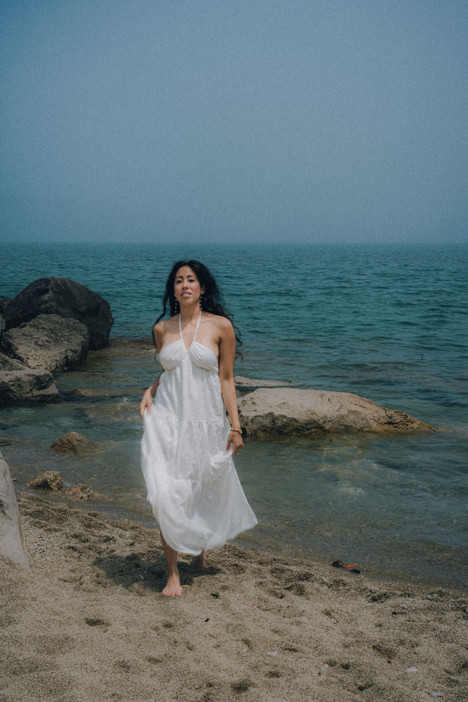 A woman in a white halter dress walks barefoot on a sandy beach with rocks and calm ocean waters in the background.