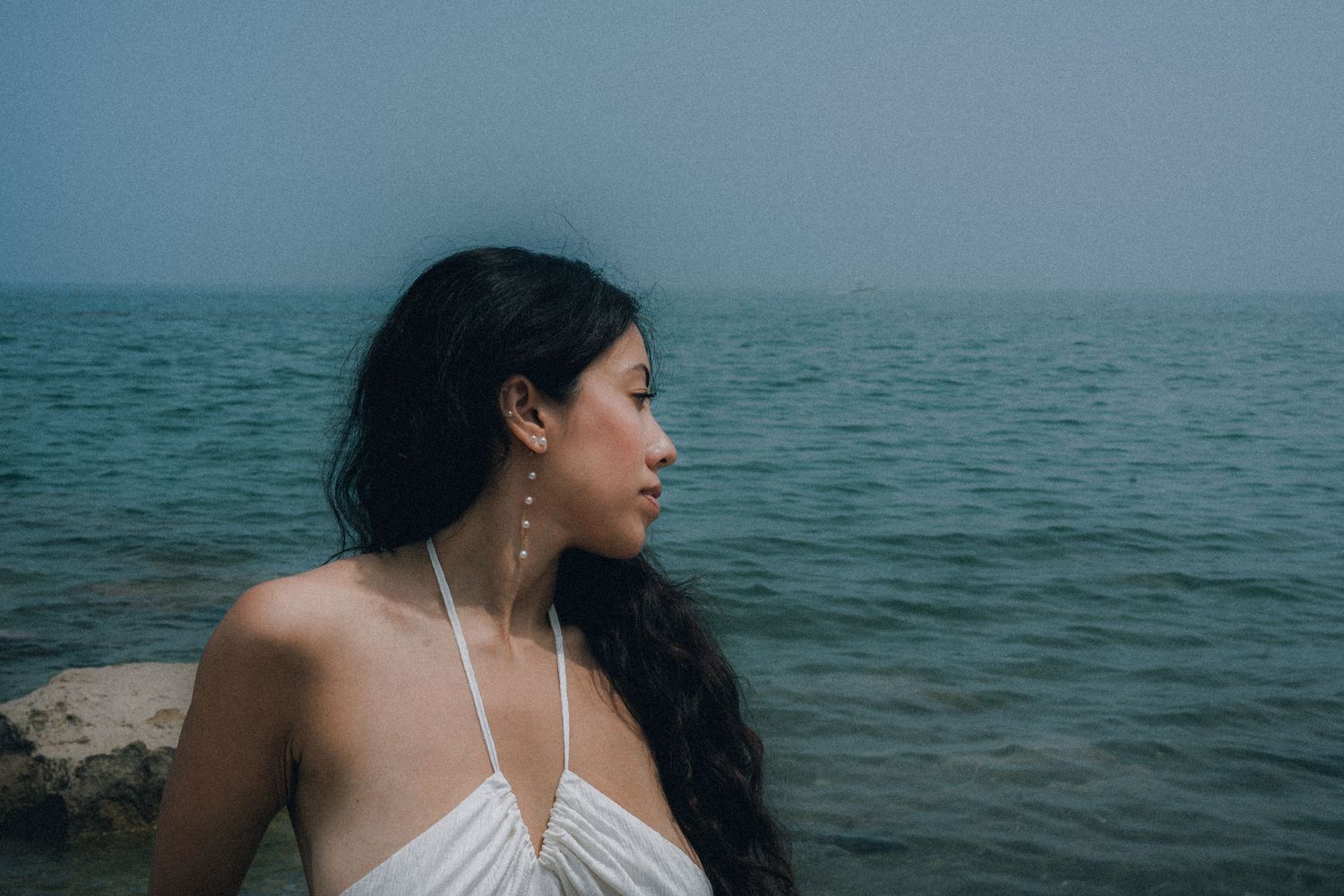 A young woman in a white dress gazes at the ocean, wearing long earrings, with misty blue water in the background.