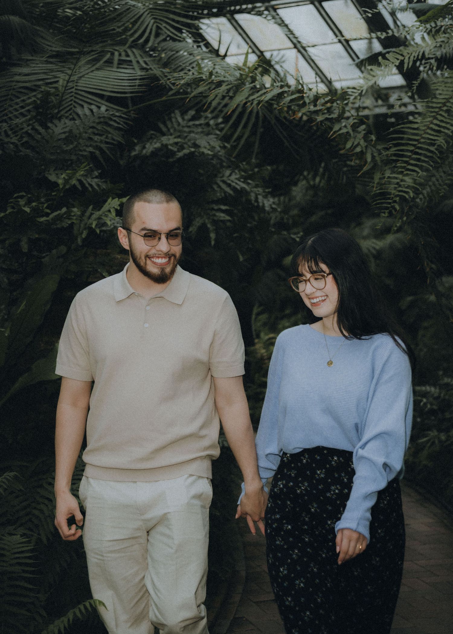 A couple stands together in a greenhouse surrounded by lush ferns and tropical plants.