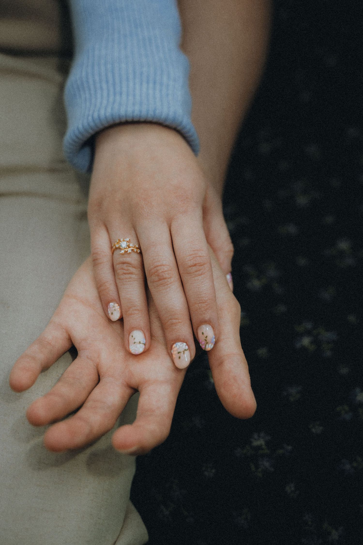 Close-up photo of hands wearing a light blue sweater showing off a diamond engagement ring on marble surface.