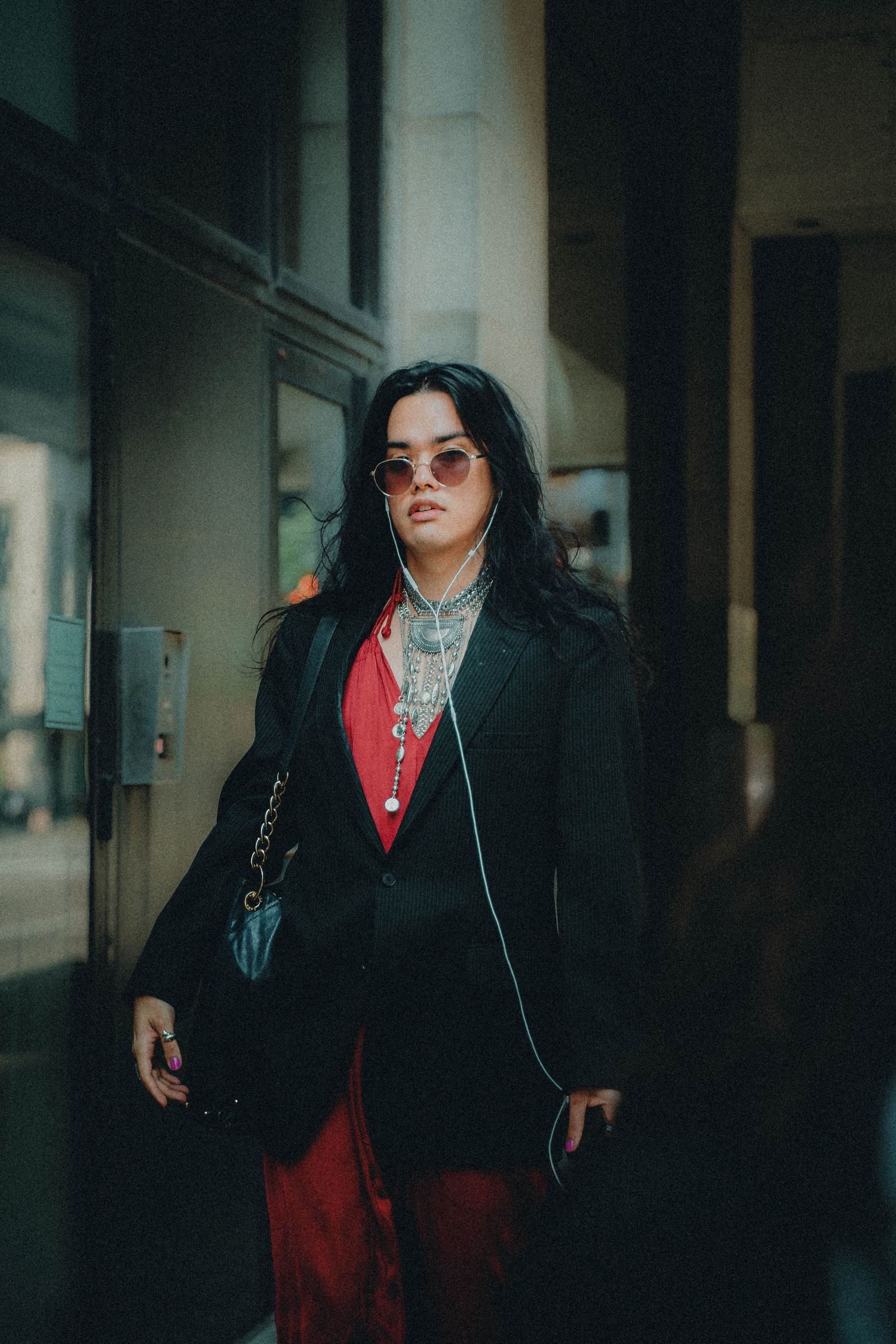 A person in a black coat and red outfit walks through a moody, dimly lit corridor with sunglasses.