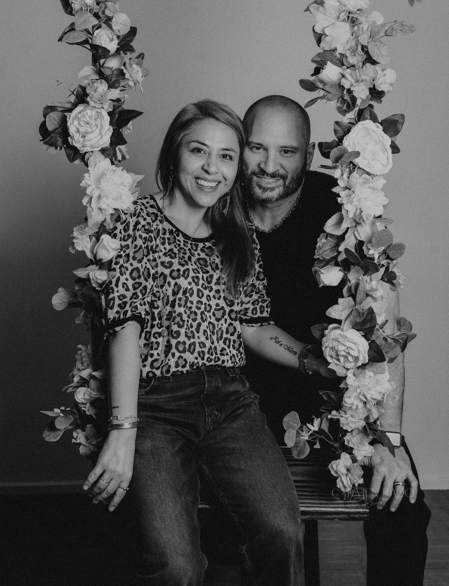 A couple sits together on a bench with decorative white floral arrangements in a casual photo shoot setting.