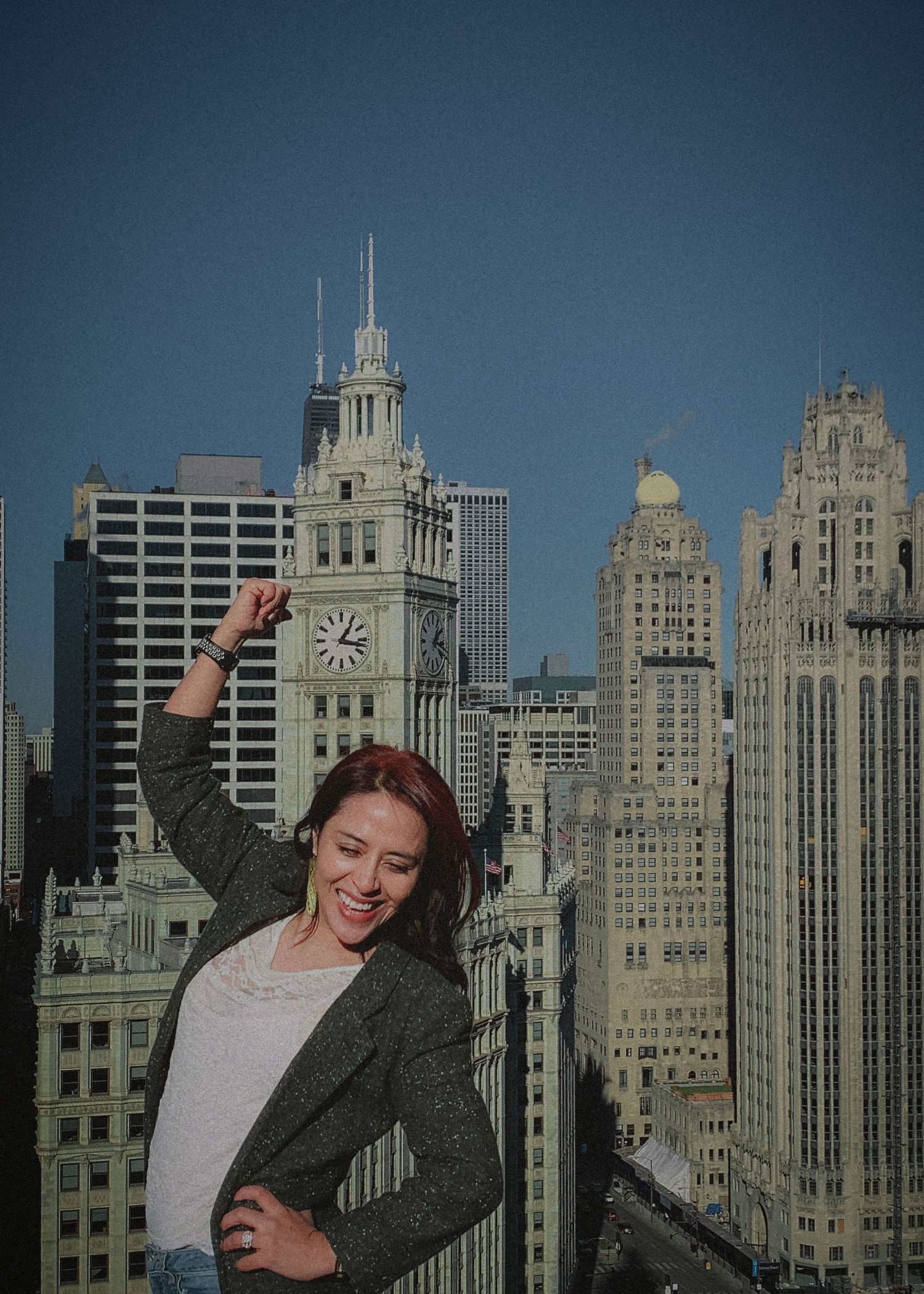 A person in a gray jacket poses playfully in front of the Chicago skyline on a sunny day.