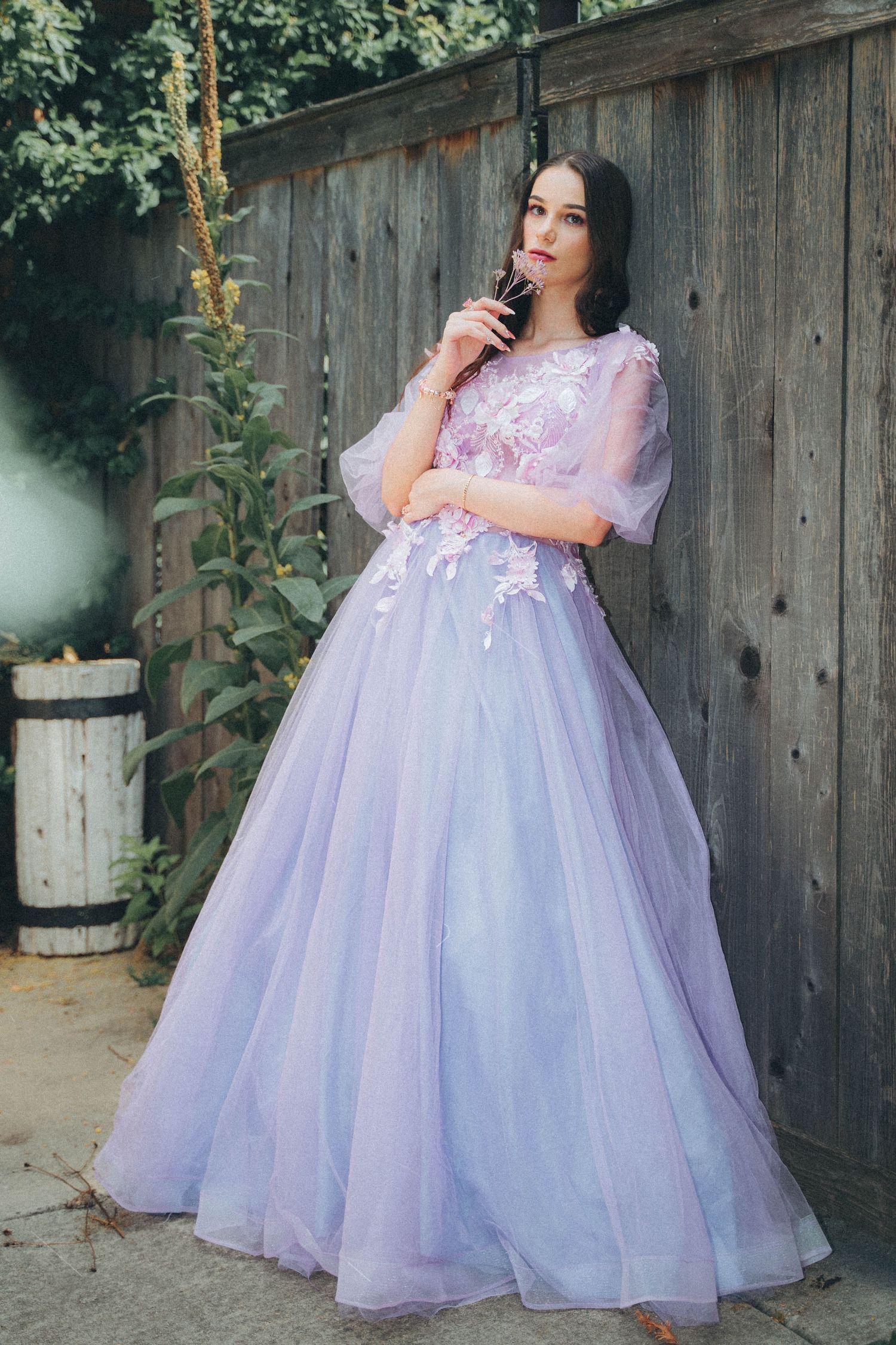 A model in a dreamy lavender tulle ball gown poses against a rustic wooden fence in an outdoor garden setting.
