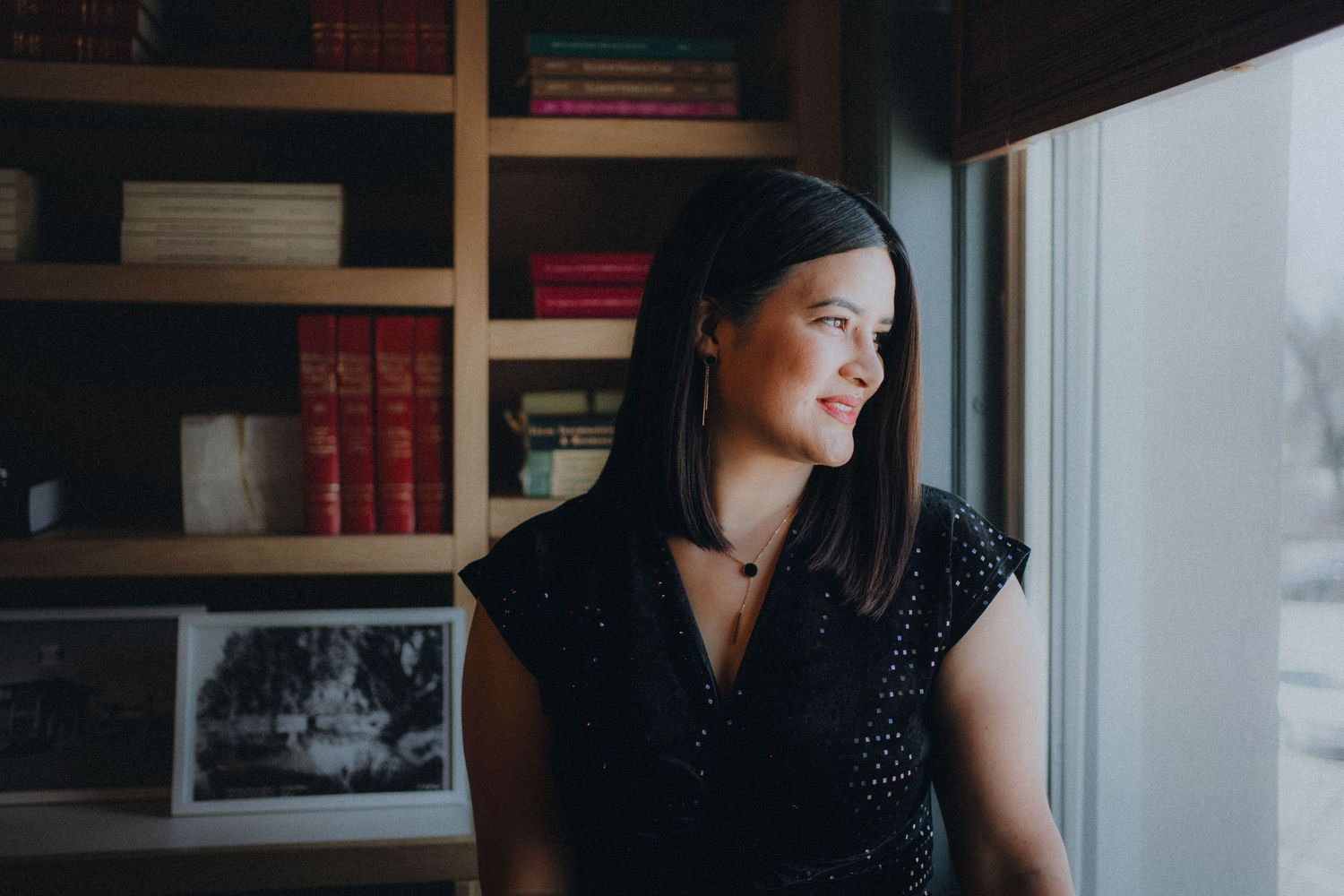 A person with dark hair sits thoughtfully by a window with bookshelves in the background.