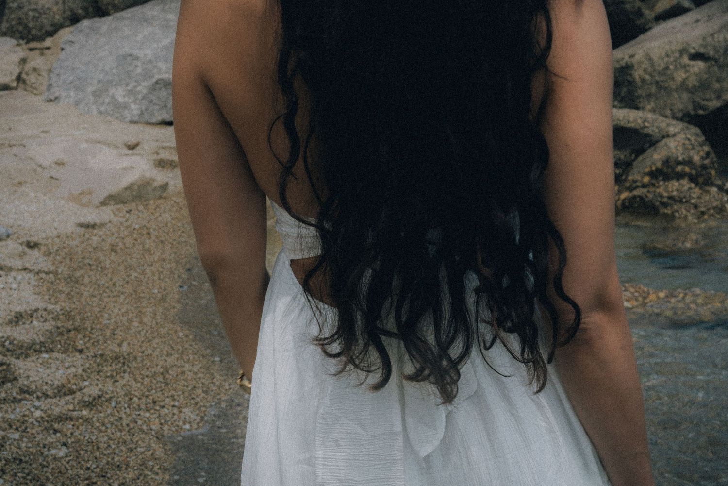 Woman in white dress with long dark curly hair standing on a rocky beach, viewed from behind.