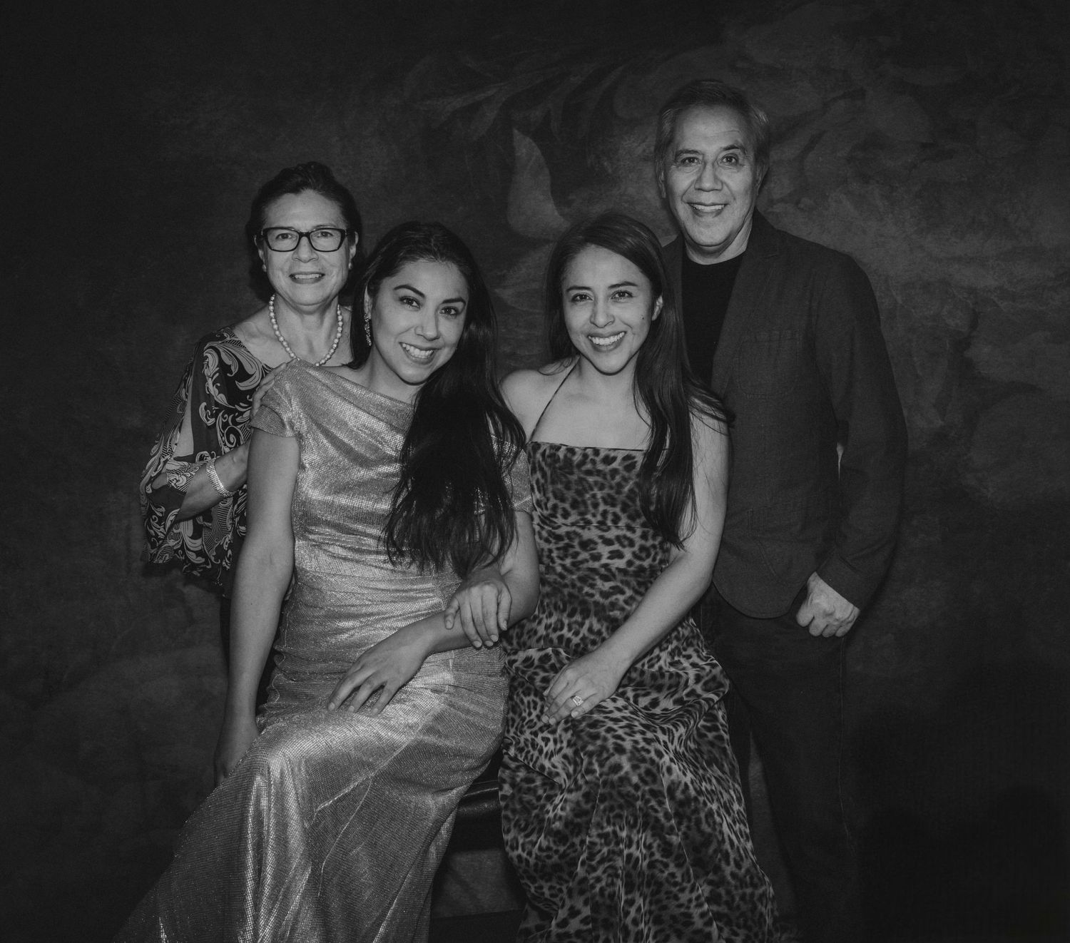 A black and white portrait of four people in formal attire posing together against a dark background.
