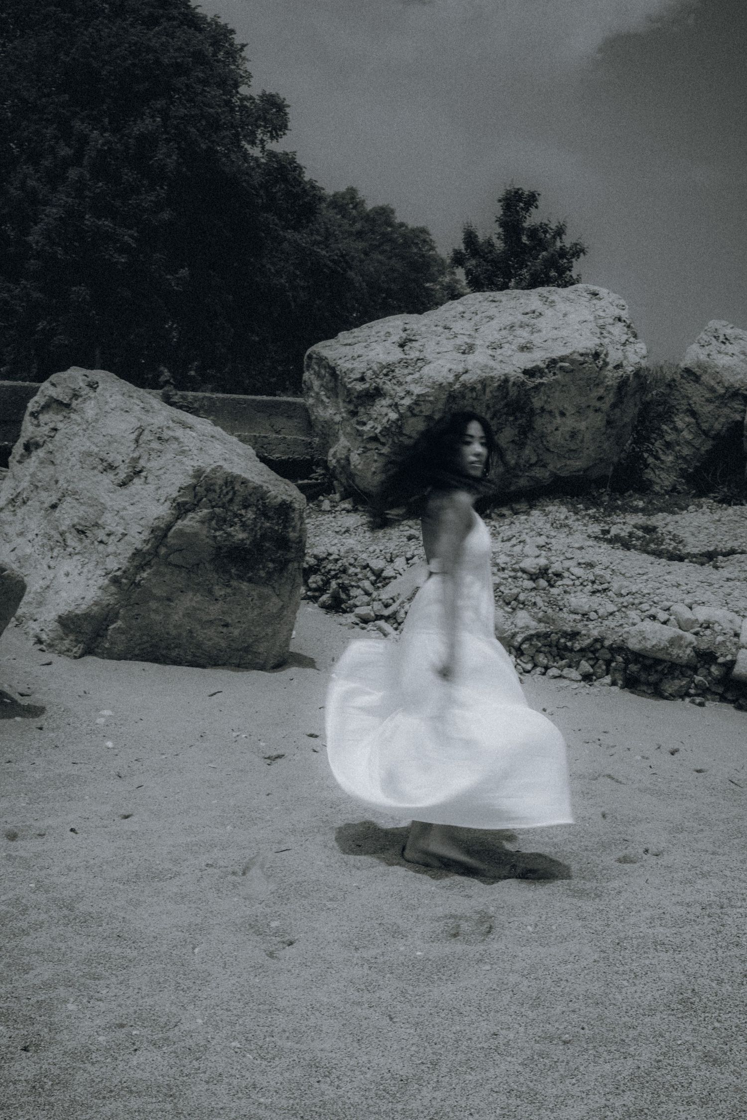 Black and white photo of a woman in a flowing white dress spinning on a sandy beach surrounded by large rocks.