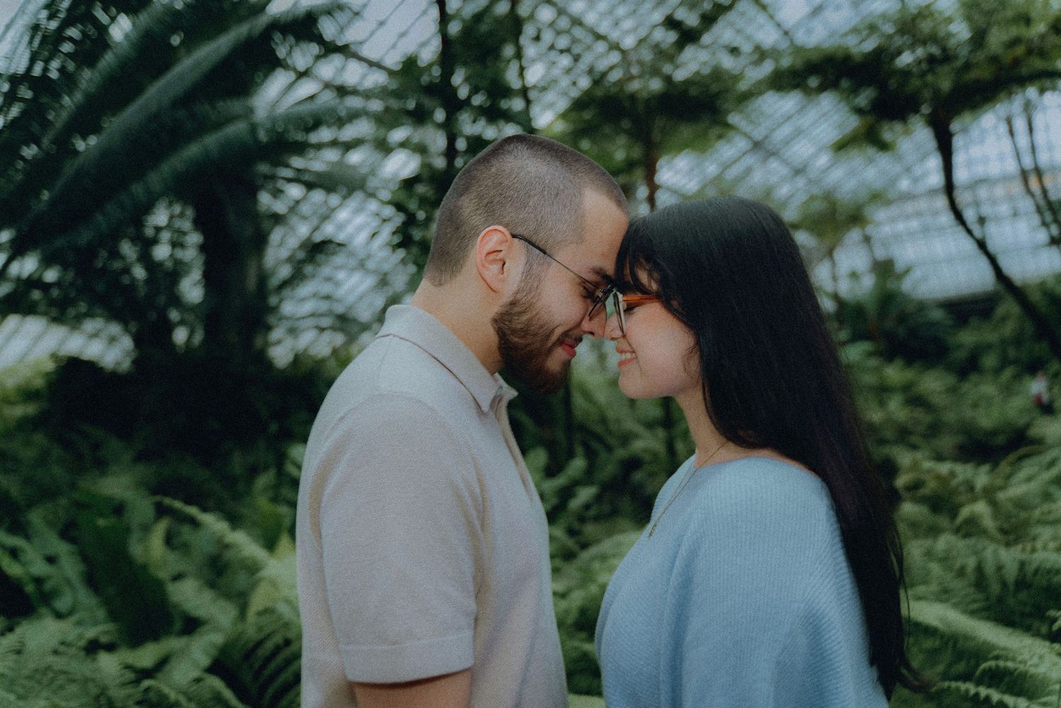 Couple sharing a romantic moment face-to-face surrounded by lush green ferns in a botanical garden.
