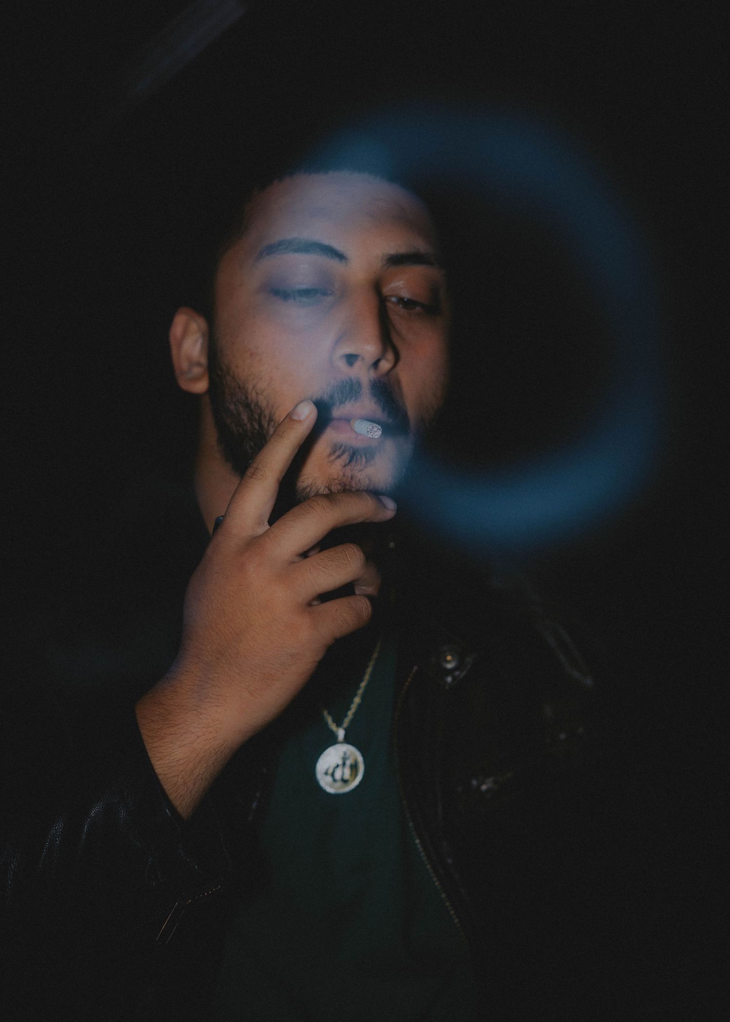 Moody portrait in dark lighting with blue halo effect shows person smoking while wearing black jacket and pendant necklace.