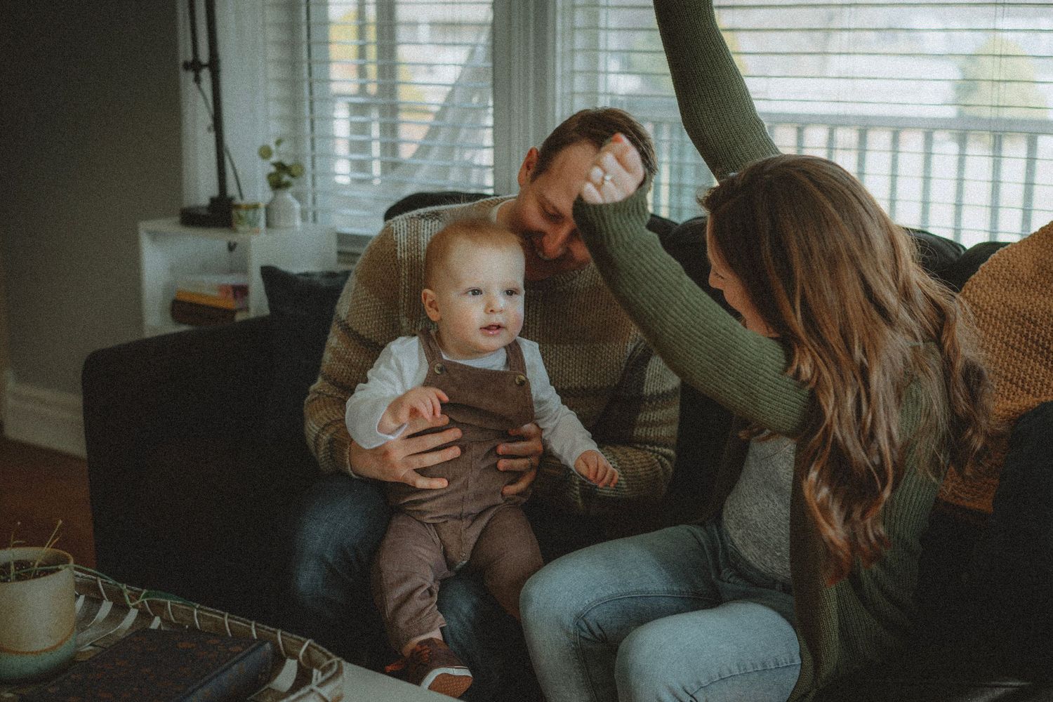 A young family sits on a couch, parents playfully interacting with their smiling baby in a cozy living room.