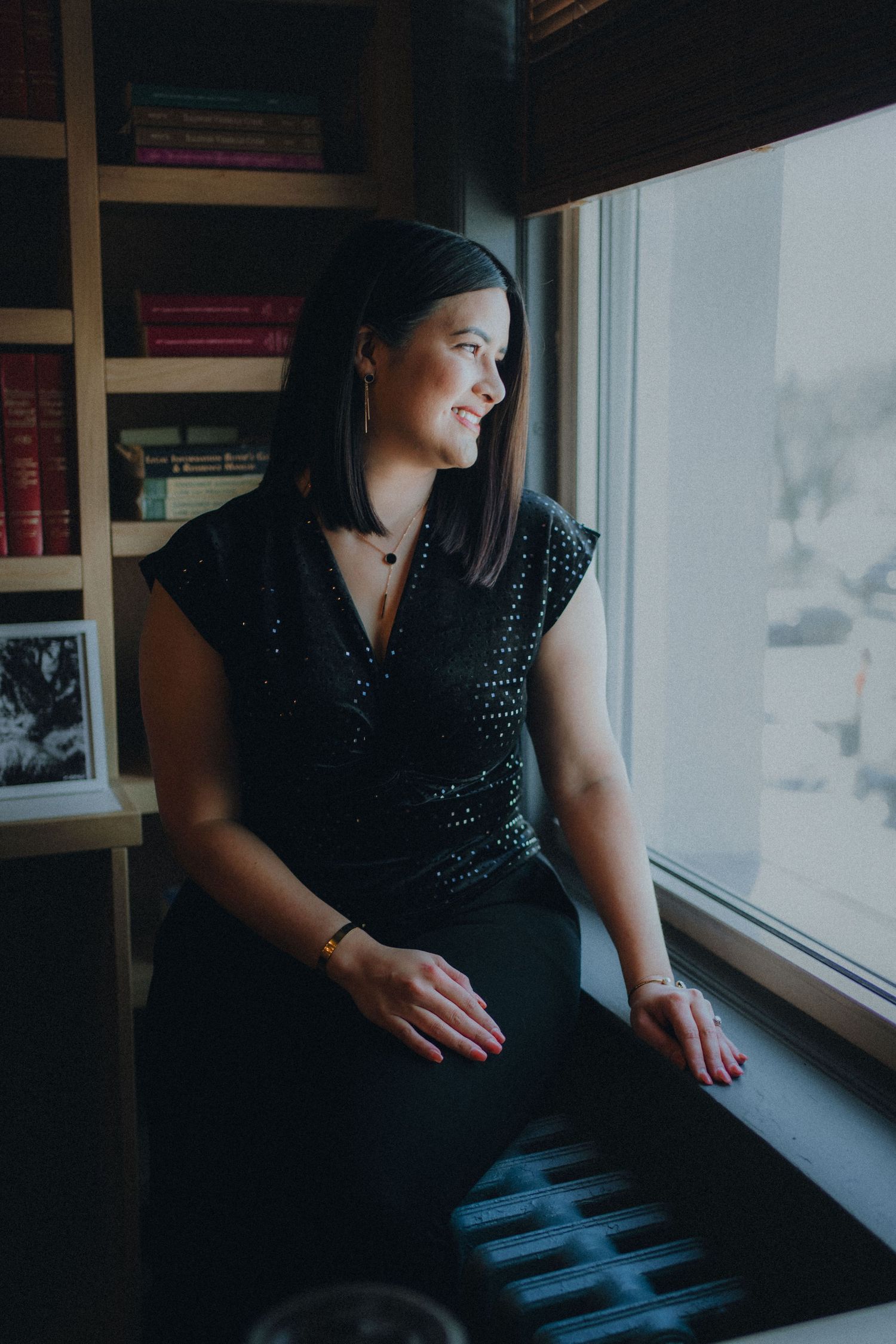 Woman in black dress sitting on windowsill, gazing outside with bookshelves in background.