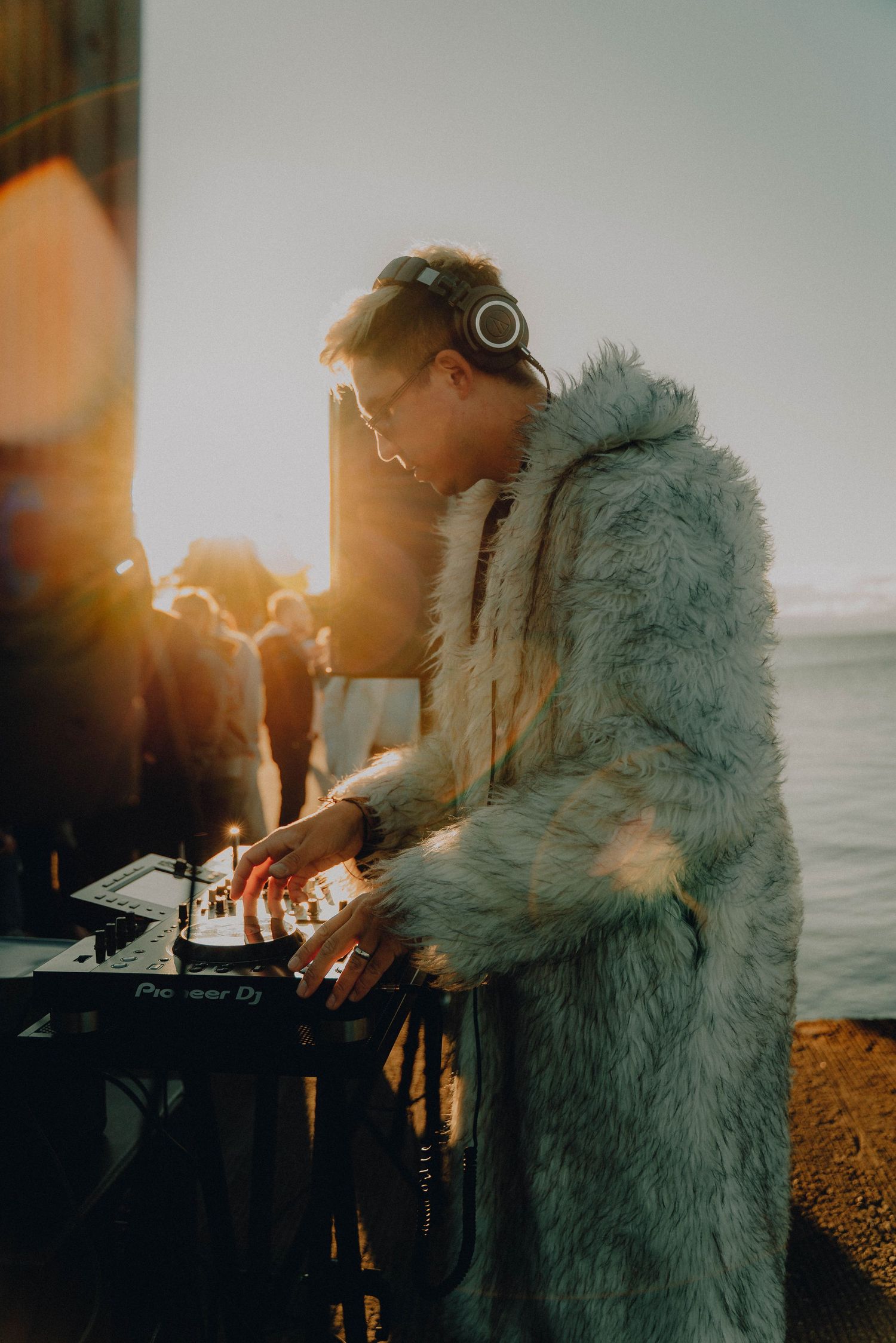 DJ performs at sunset beach party wearing fuzzy teal coat and headphones while mixing on turntables.