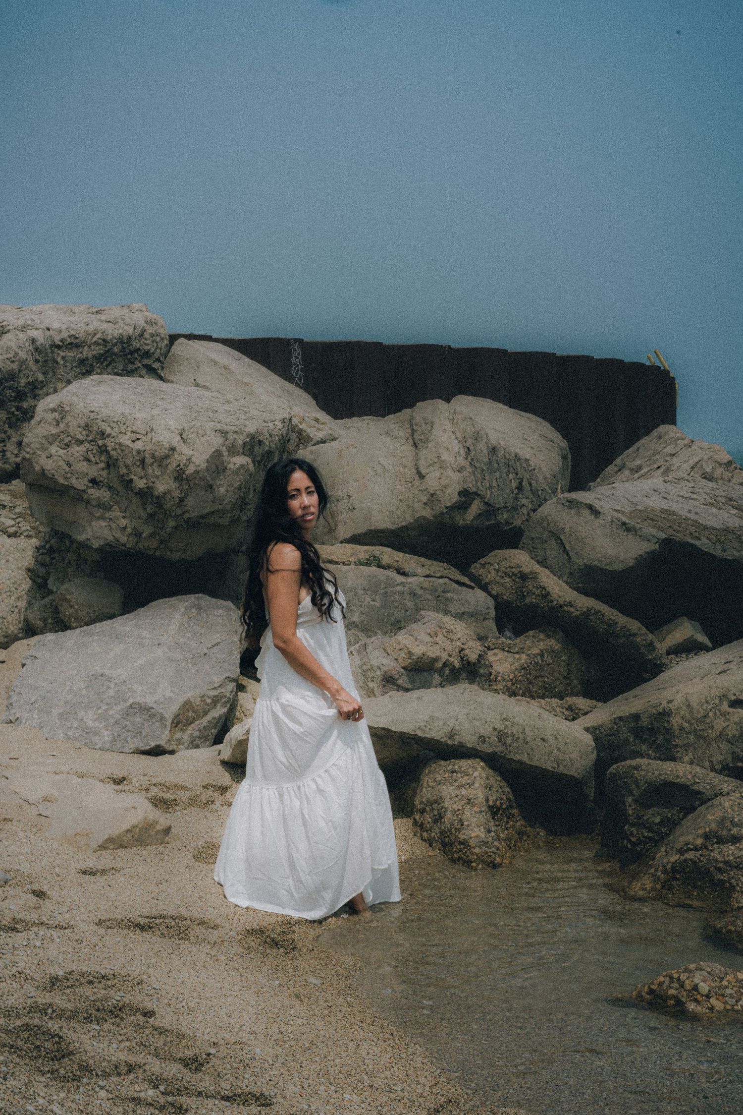 Woman in white dress standing on sandy beach among large rocks near water.