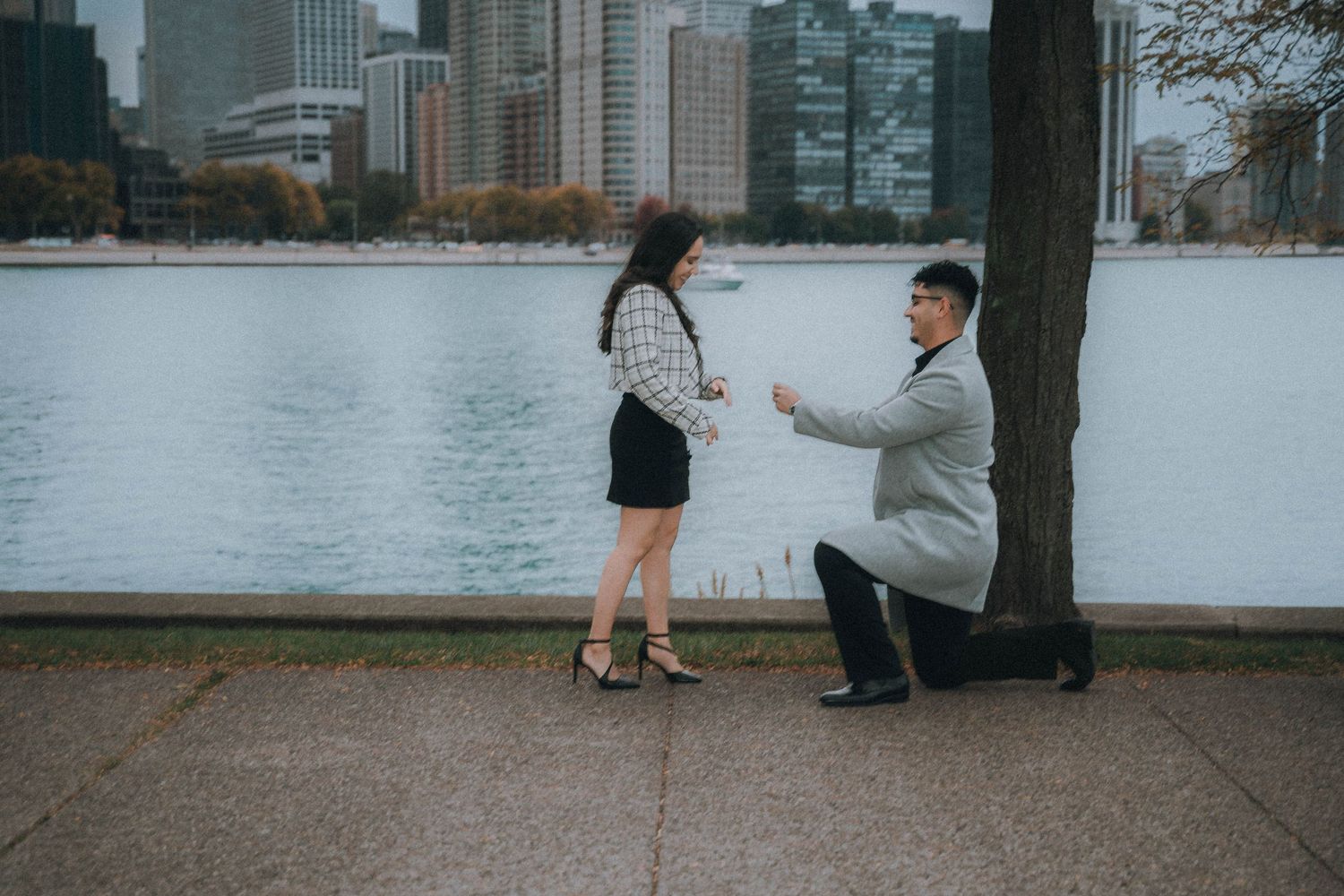 Couple holds hands while walking along an urban waterfront with city buildings in the background.