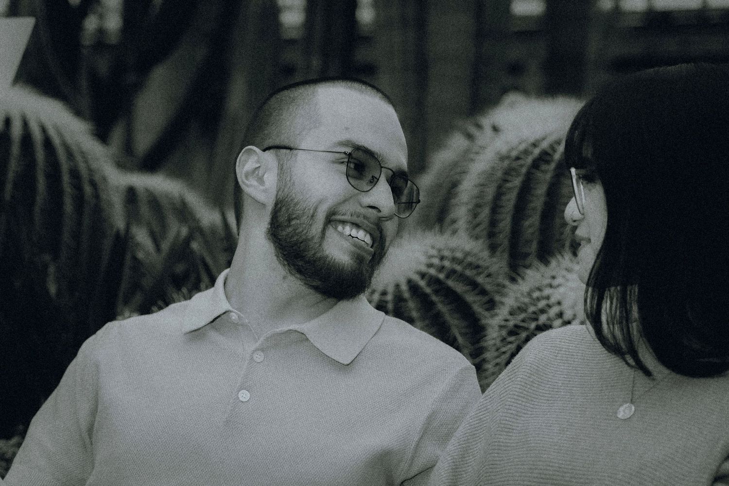 Black and white candid photo of a smiling person wearing glasses and a polo shirt during a conversation outdoors.