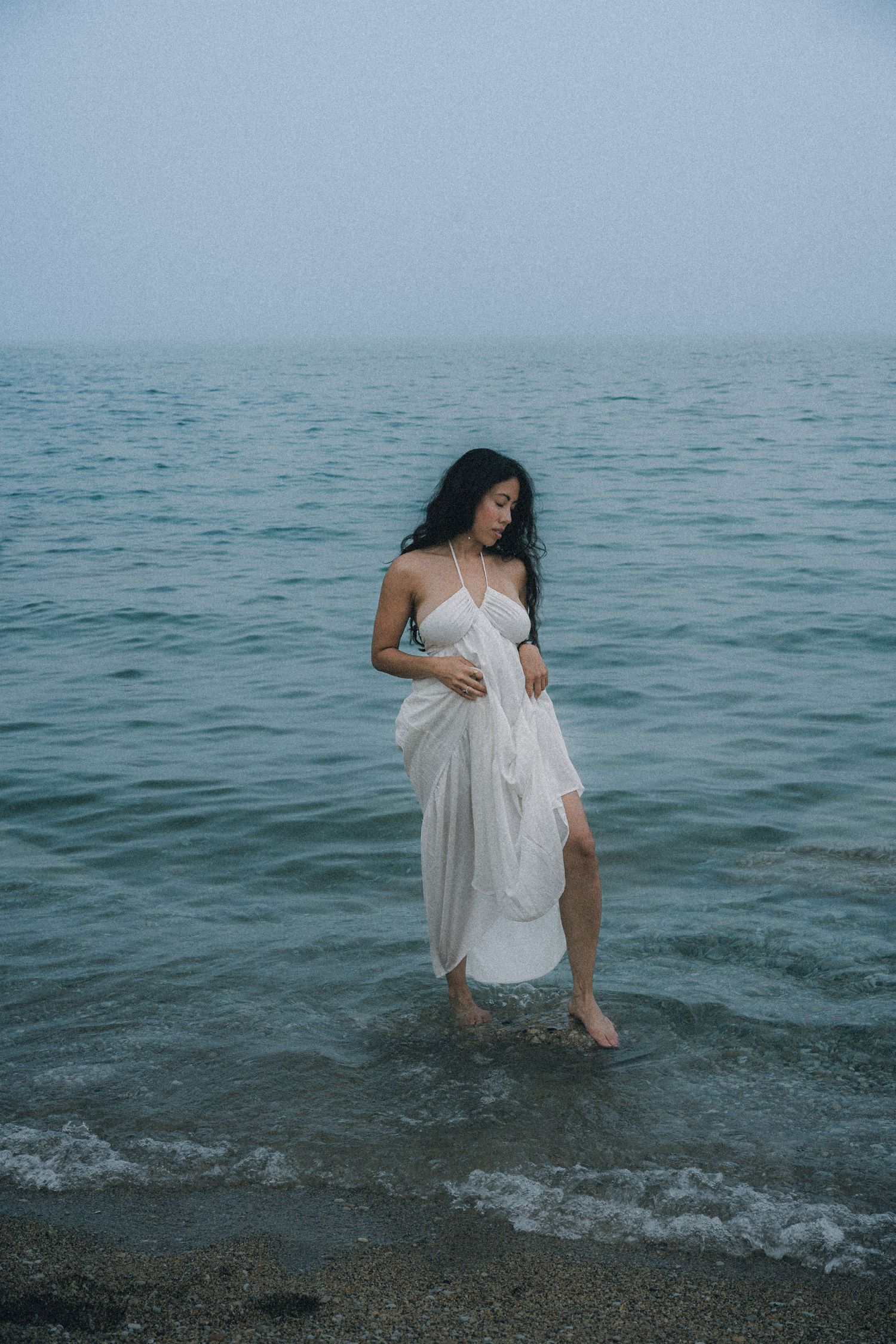 A woman in a flowing white dress stands in shallow ocean waves on a misty, overcast day.
