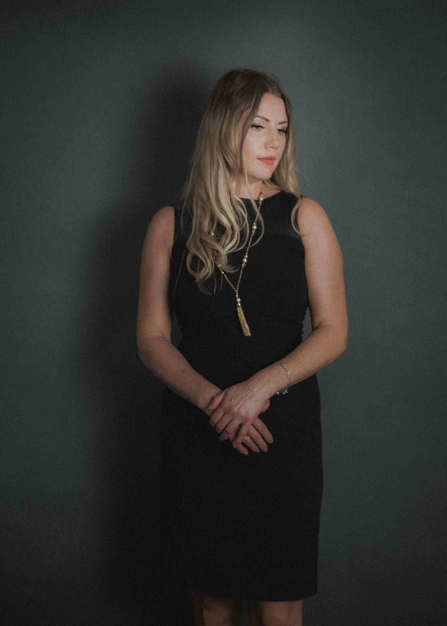 Moody indoor portrait features figure in sleek black sleeveless dress against dark green wall with dramatic lighting.