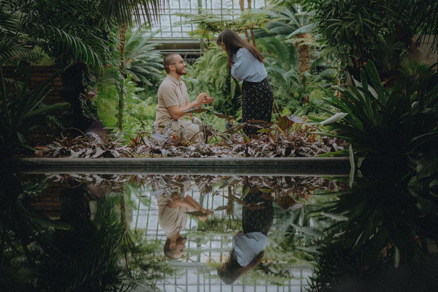 Two people share an intimate moment in a lush greenhouse with a reflective pool creating a mirror image below them.