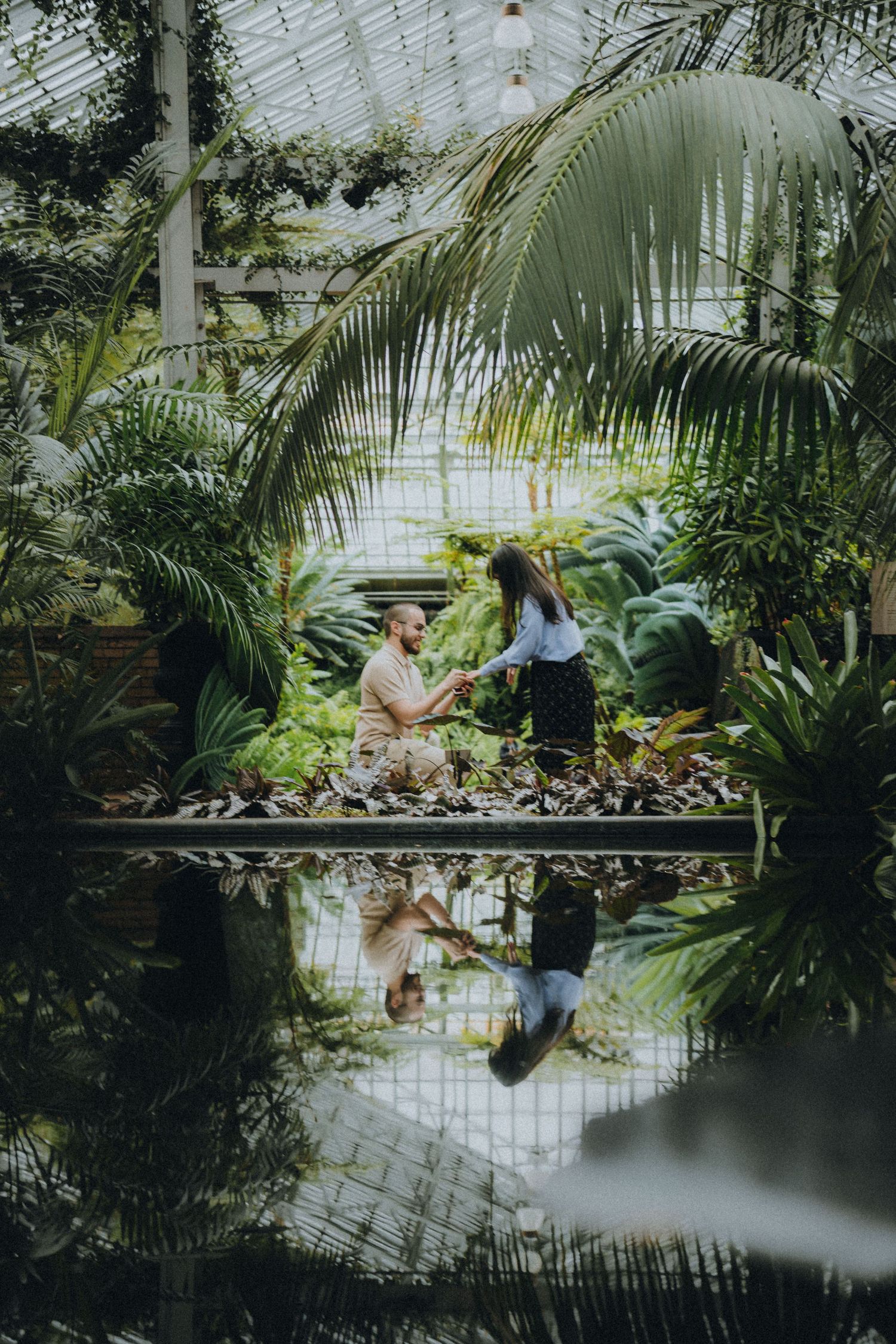 A couple sits on a bench surrounded by lush tropical plants and palm trees inside a bright greenhouse conservatory.