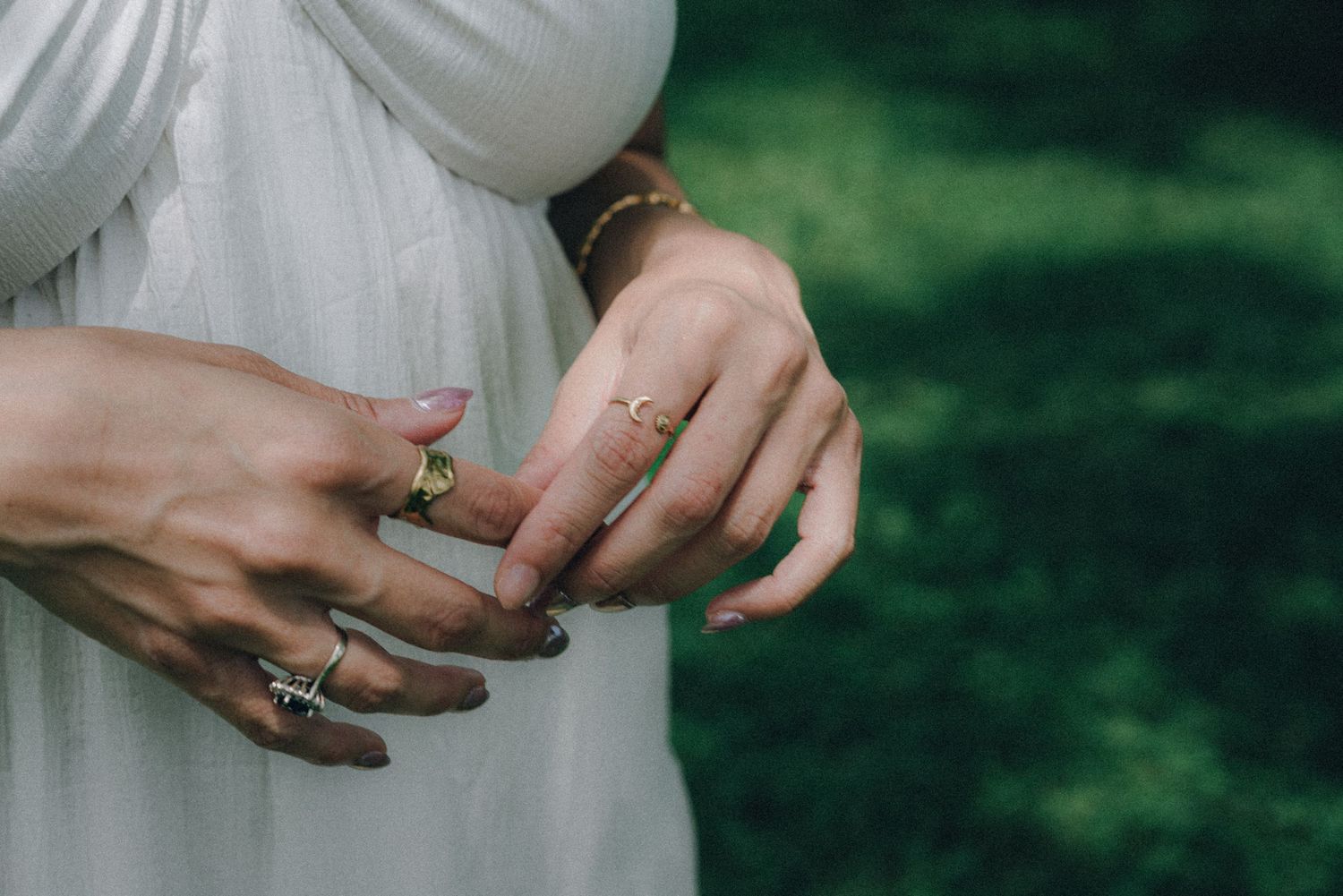 Close-up of woman's hands adorned with multiple gold and silver rings and a delicate bracelet, wearing a white dress outdoors.