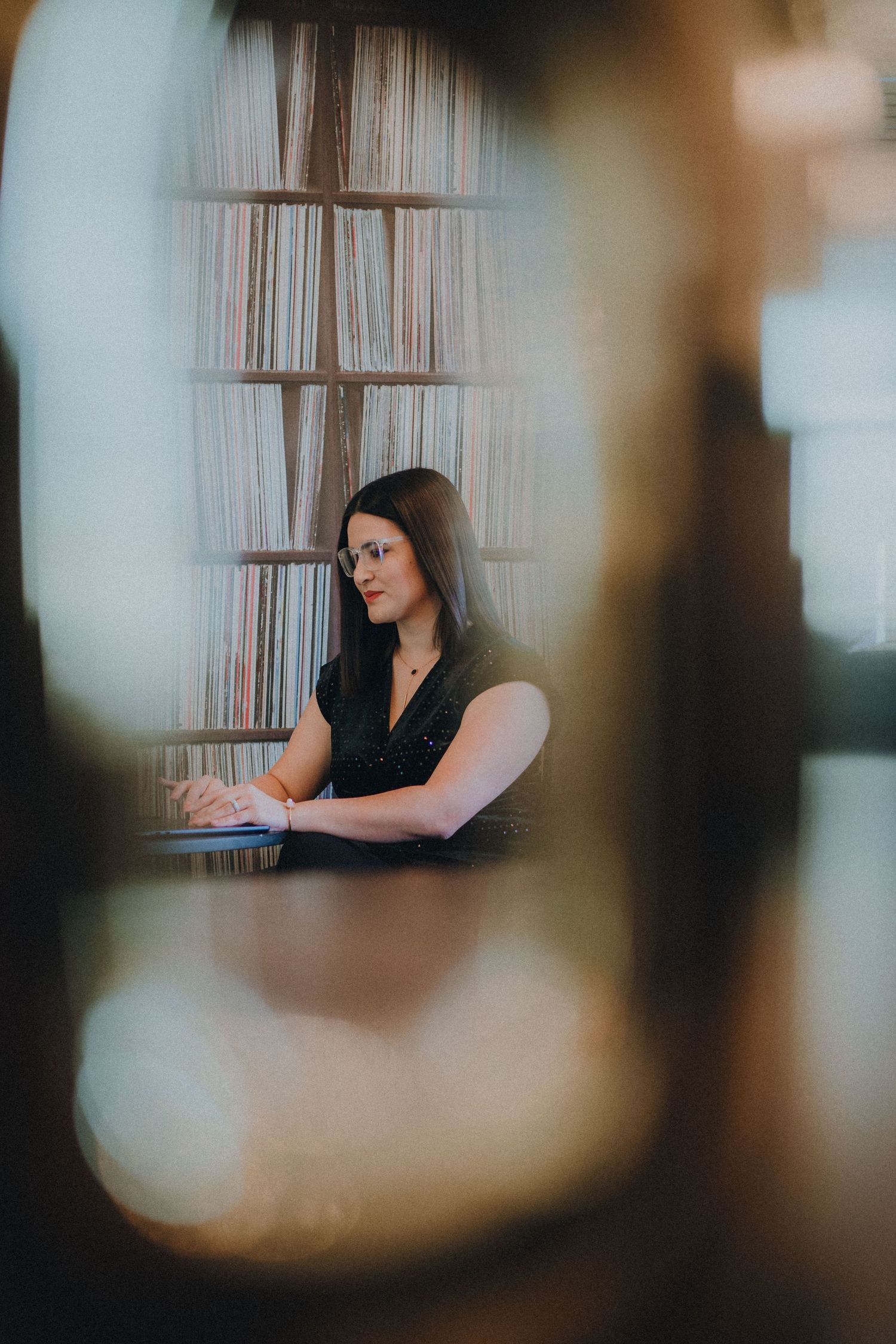 Artistic portrait series showing a seated figure in a black top against blurred foreground elements and slatted window backdrop.