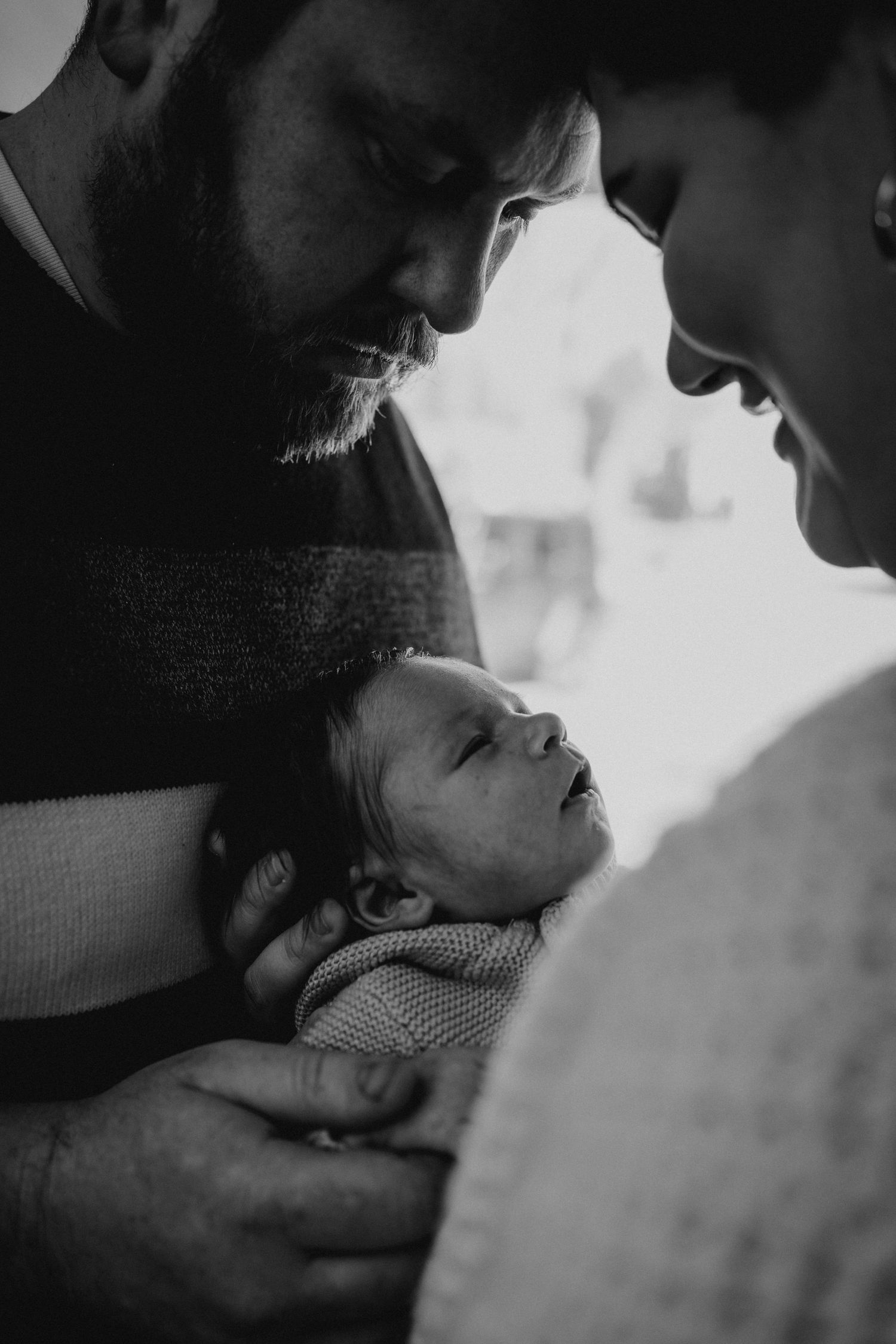 A tender black and white portrait of parents gazing lovingly at their newborn baby in an intimate moment.