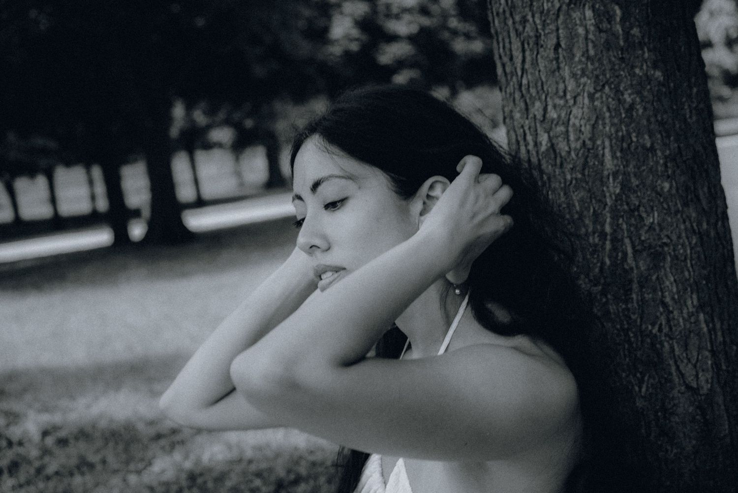 Black and white photo of a young woman leaning against a tree in a park, eyes closed, hands behind her head.