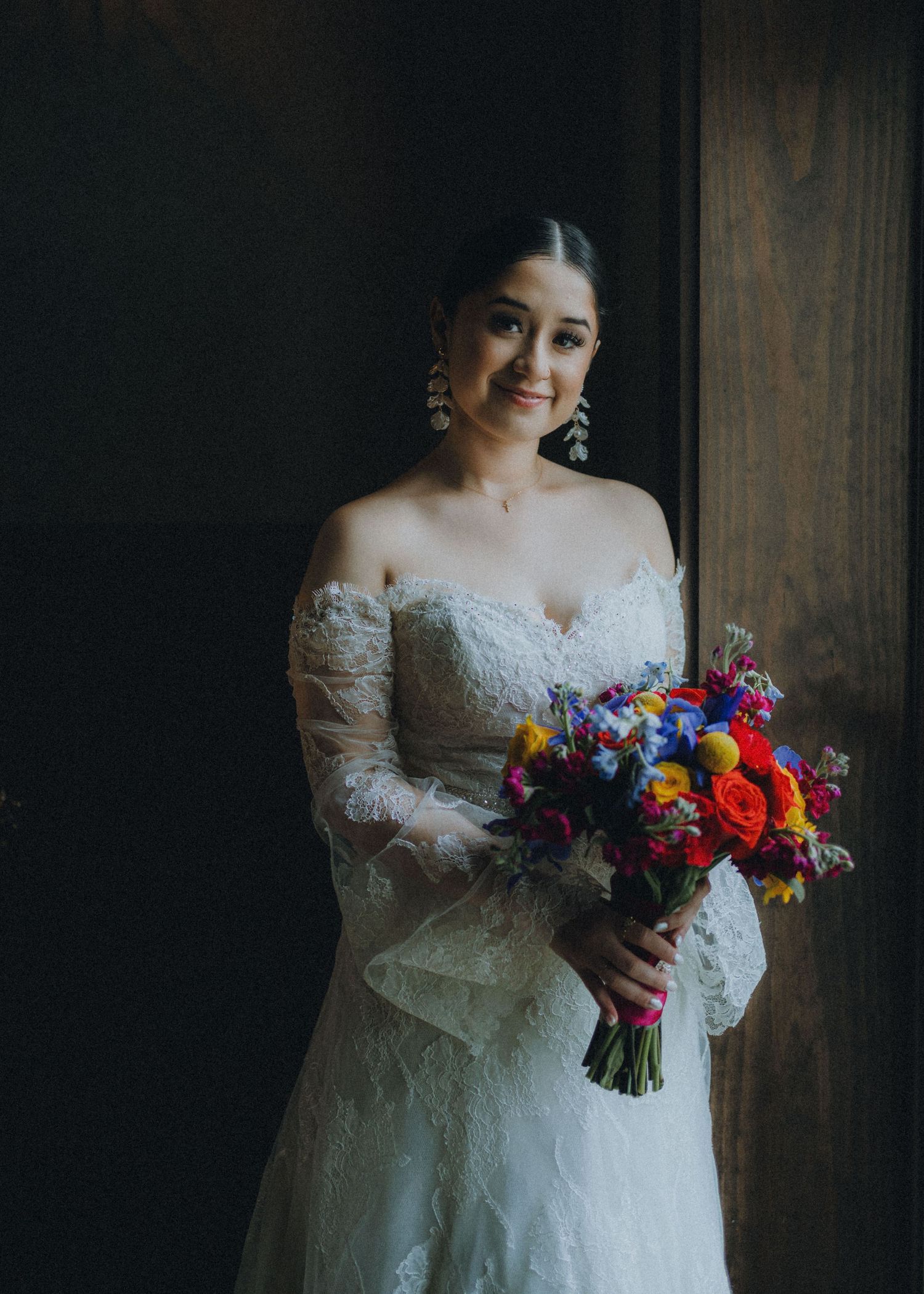 A bride in a white lace gown holds a vibrant bouquet of red, purple, and yellow flowers against a dark wooden backdrop.