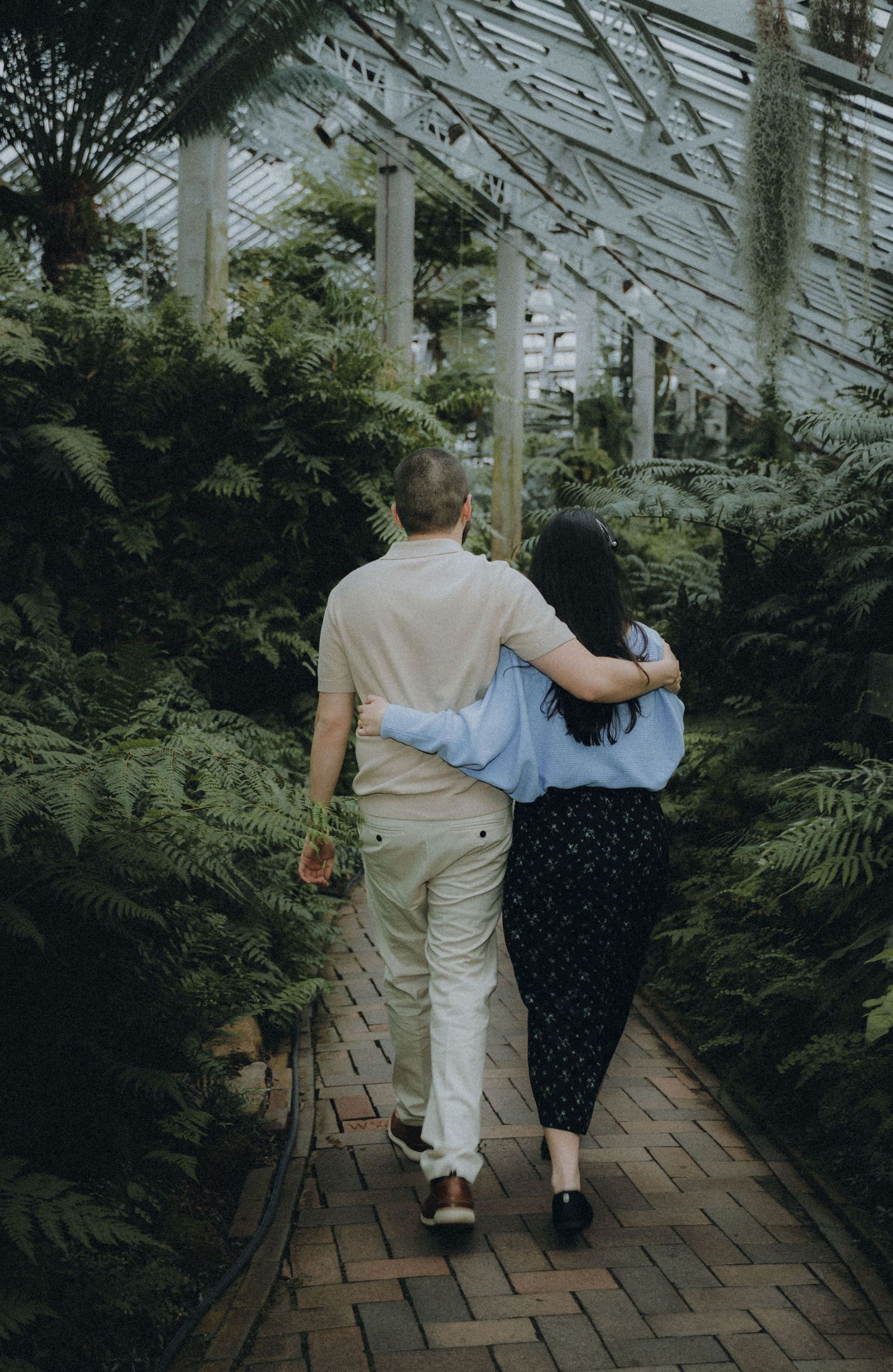 Two people walk together down a brick path in a lush greenhouse with ferns and greenery.