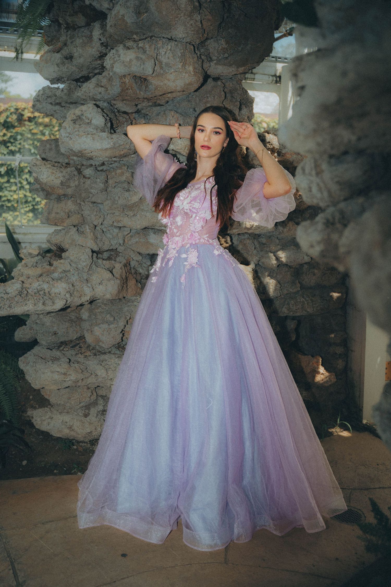 A person in a lavender tulle ball gown poses against a rustic stone wall background in a romantic setting.