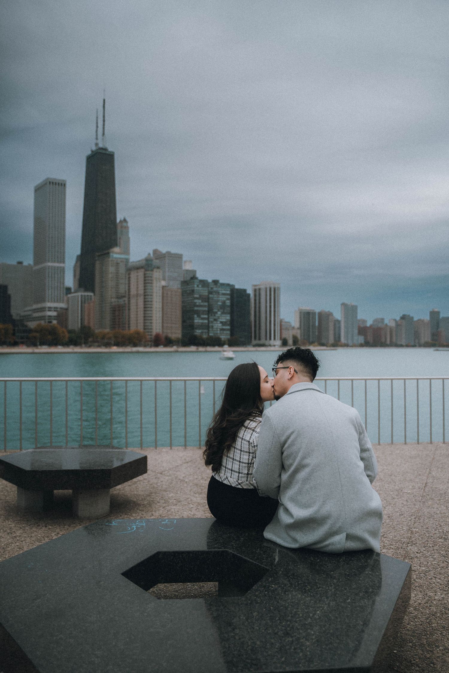 Couple kissing on a bench with Chicago skyline and Lake Michigan in the background on a cloudy day.