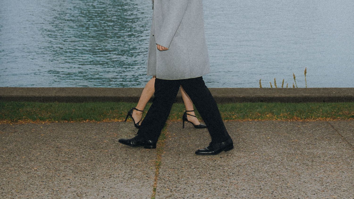 Feet of people walking on a gray concrete surface near water's edge.