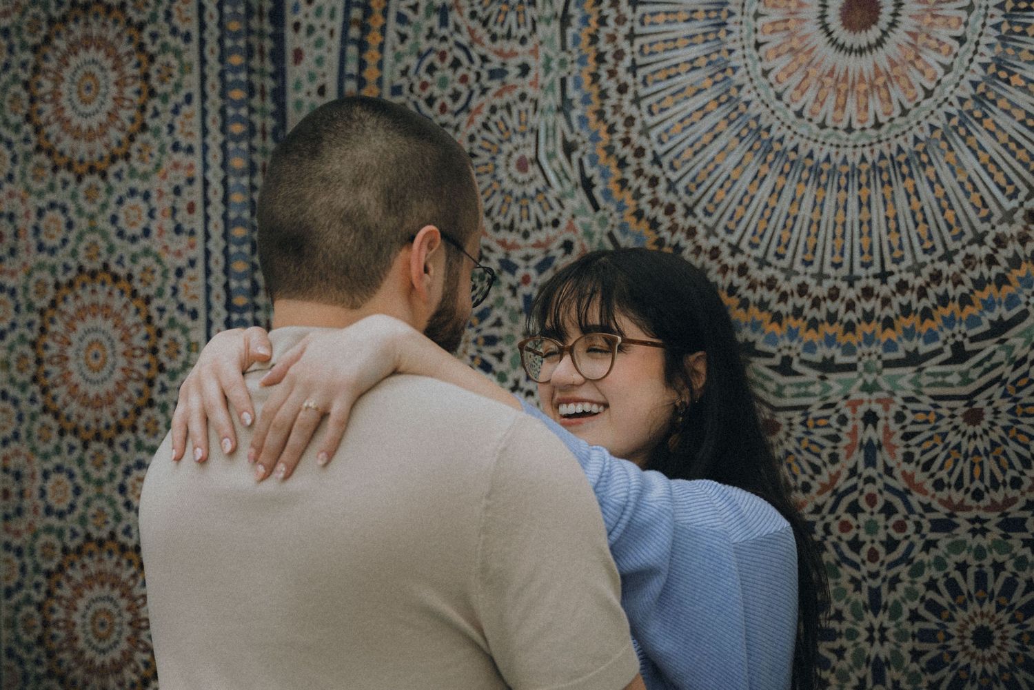 Two people embrace in front of an ornate Moroccan mosaic wall with intricate geometric patterns.