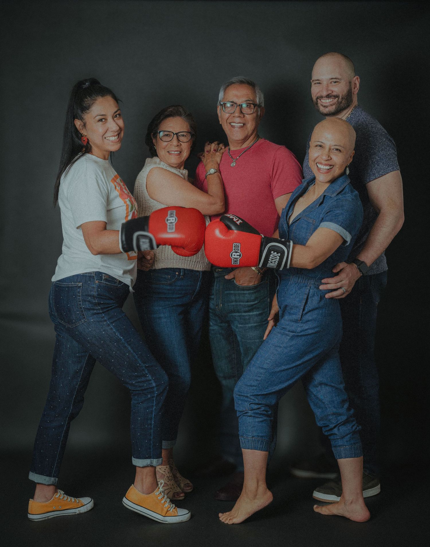 A group of four people wearing casual denim outfits pose together while holding red boxing gloves against a dark background.