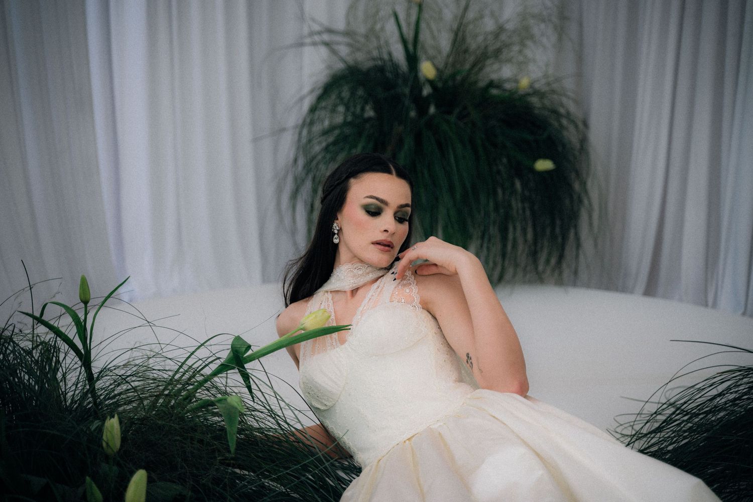 A bride in a flowing white wedding dress poses elegantly against a light curtained backdrop with green plants.