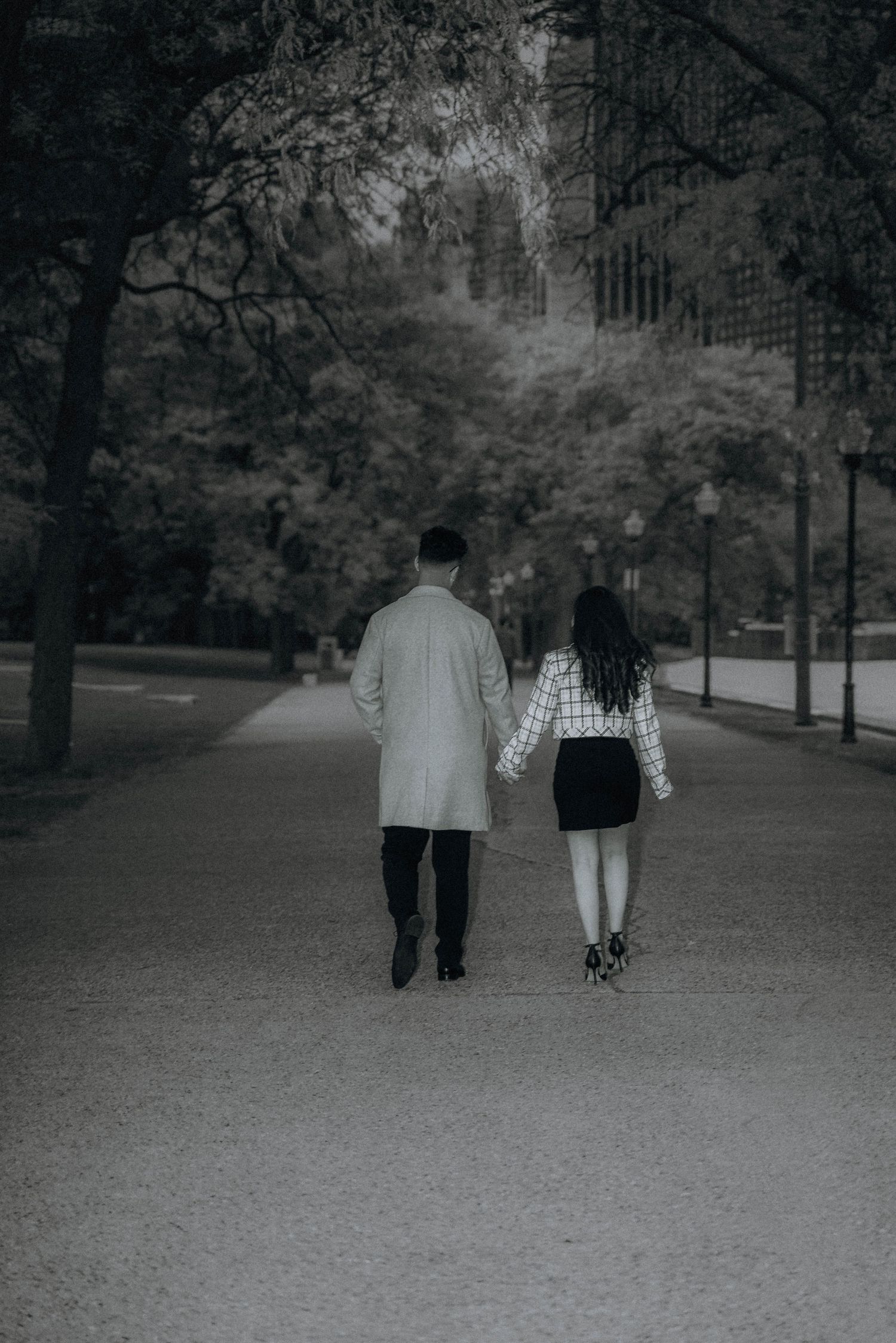 Couple holding hands walking through a tree-lined park path in black and white photography.