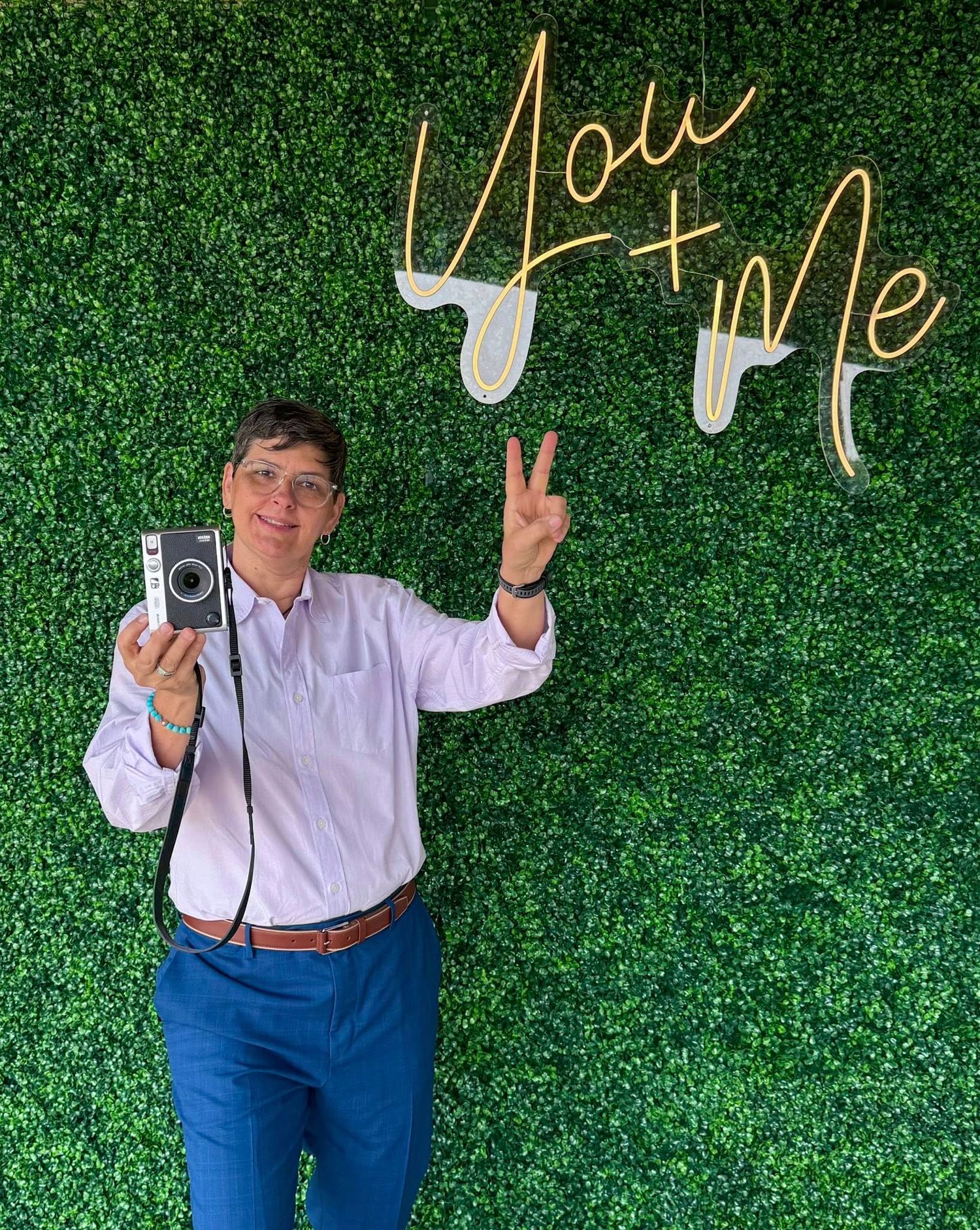 Someone poses with a vintage camera making a peace sign against a green hedge wall with 