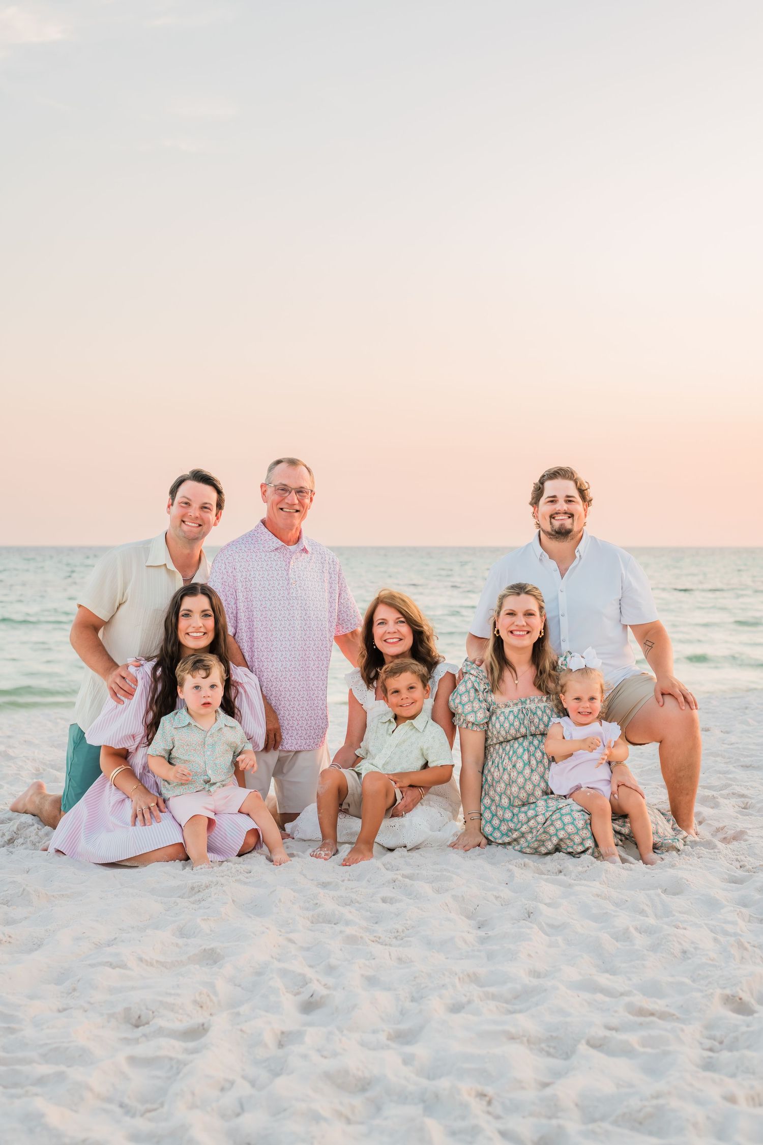 A family group wearing coordinated pastel and white outfits poses together on a sandy beach at sunset.