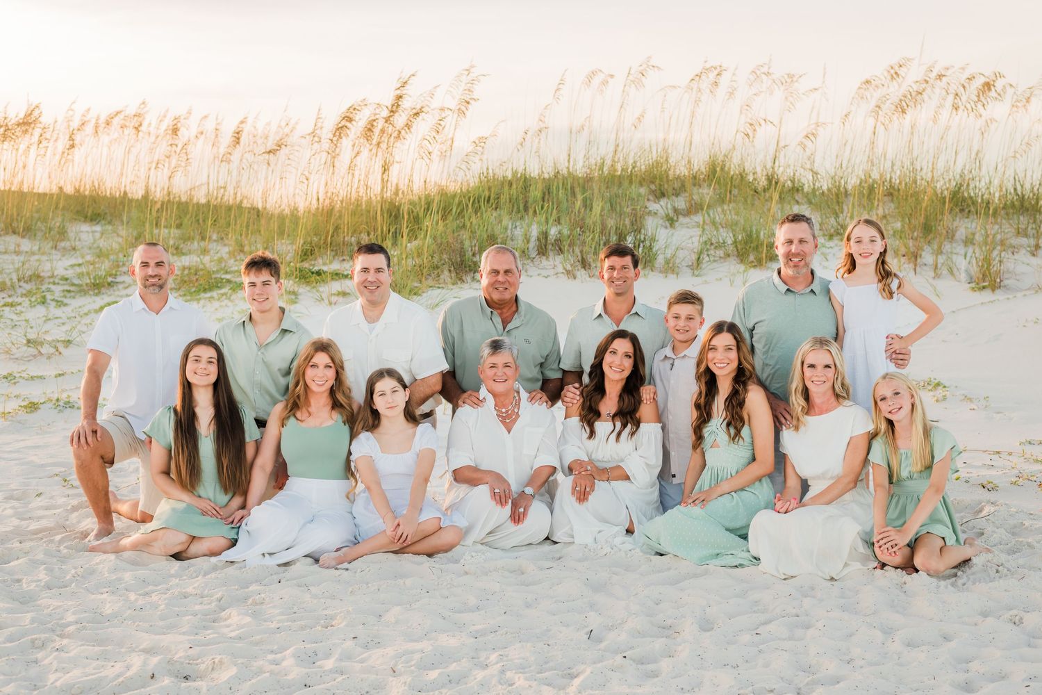 A group wearing coordinated pastel outfits poses for family portraits on a sandy beach at sunset.